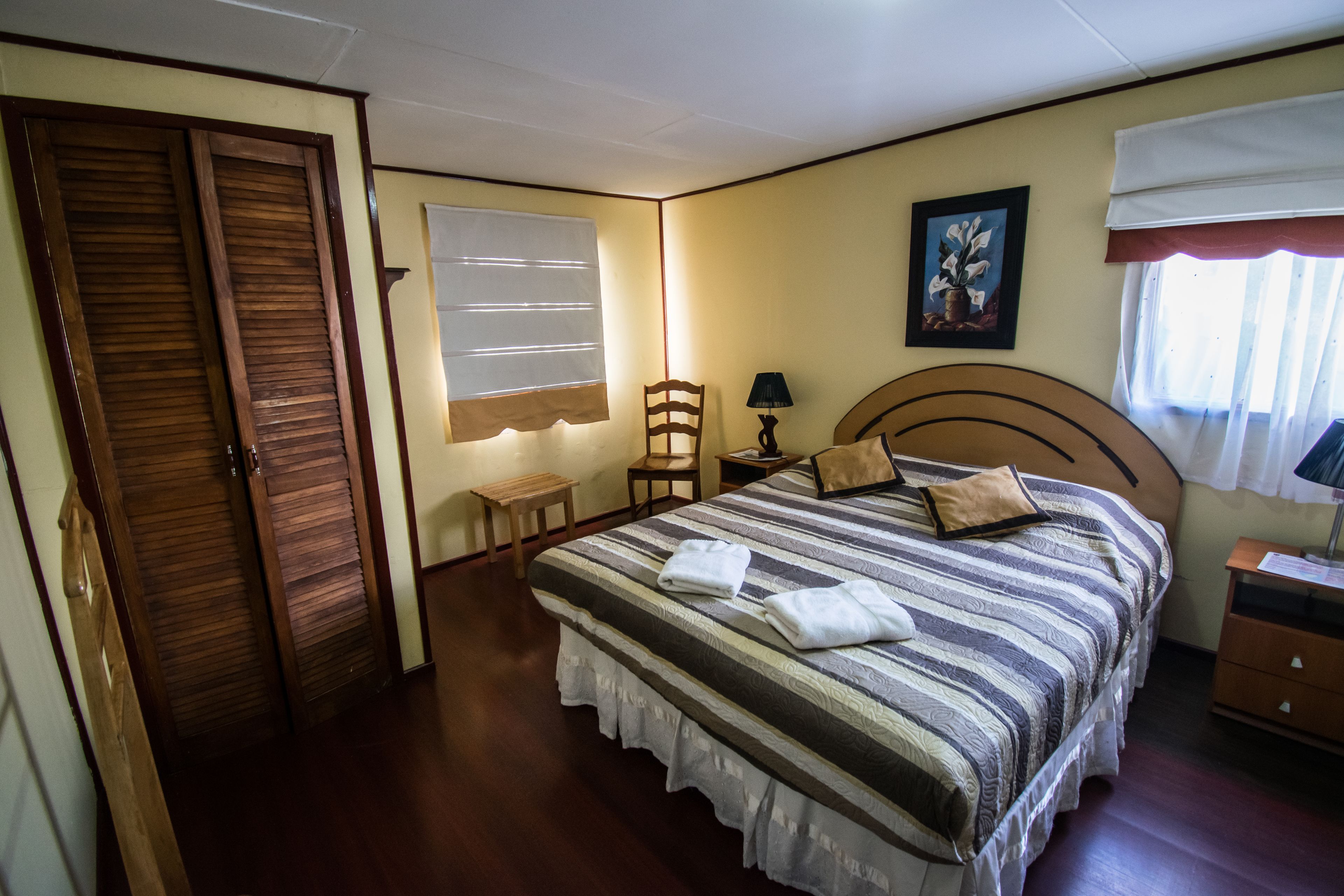A bedroom with a large bed, a wooden headboard, a nightstand, a chair, a lamp, and a window with curtains.