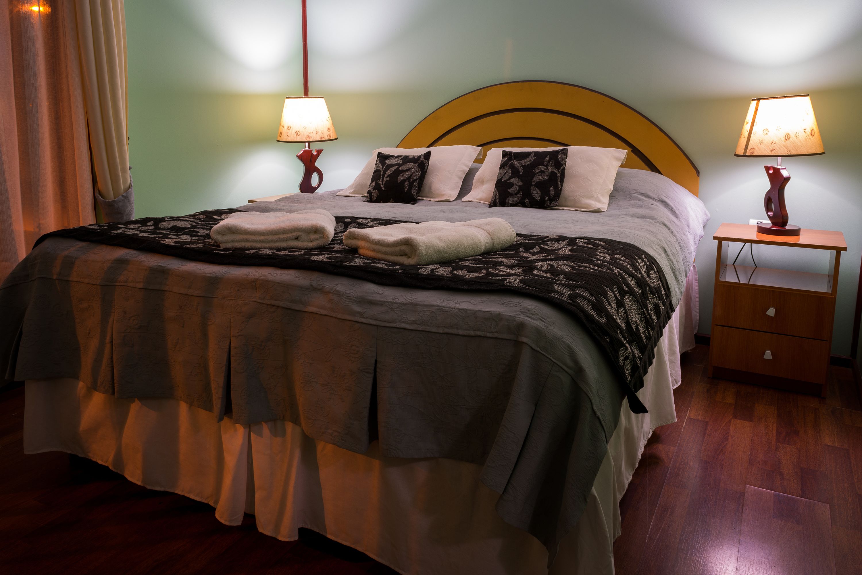 Una cama bien hecha con una colcha estampada, dos lámparas de noche y una mesita de noche de madera con cajones.