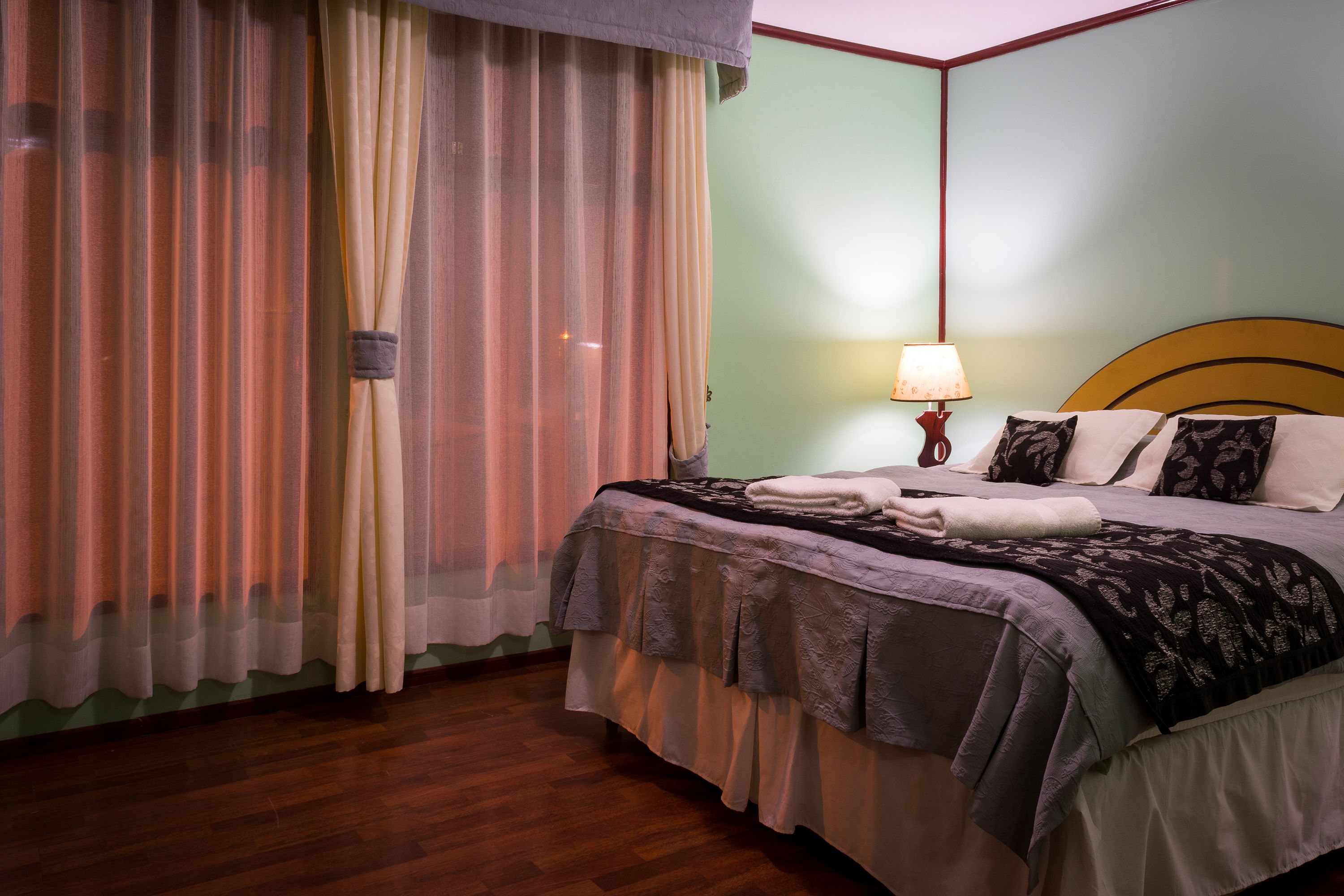 A neatly made bed with a headboard, a bedside lamp, and a window with sheer curtains.