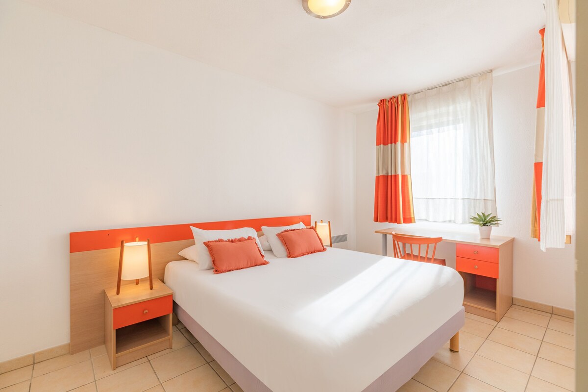 Habitación de hotel con una cama grande, una mesita de noche con lámpara, un escritorio con silla y una ventana con cortinas.