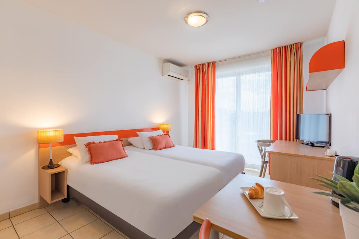 Een hotelkamer met twee bedden, een bureau met televisie, een lamp en een ontbijttafel met croissant en koffie.