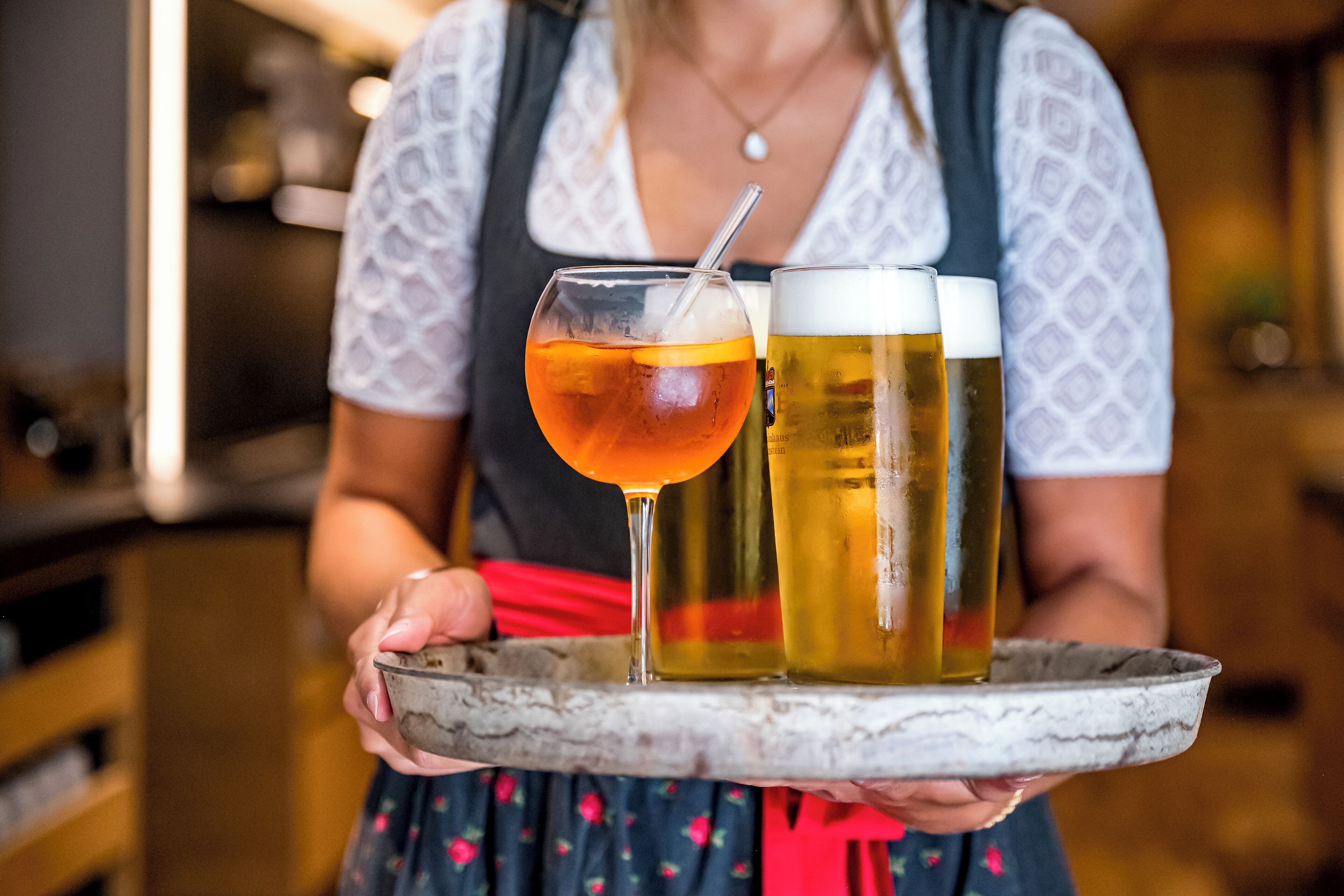 Eine Person hält ein Tablett mit einem Glas bernsteinfarbenen Getränks und zwei Biergläsern.