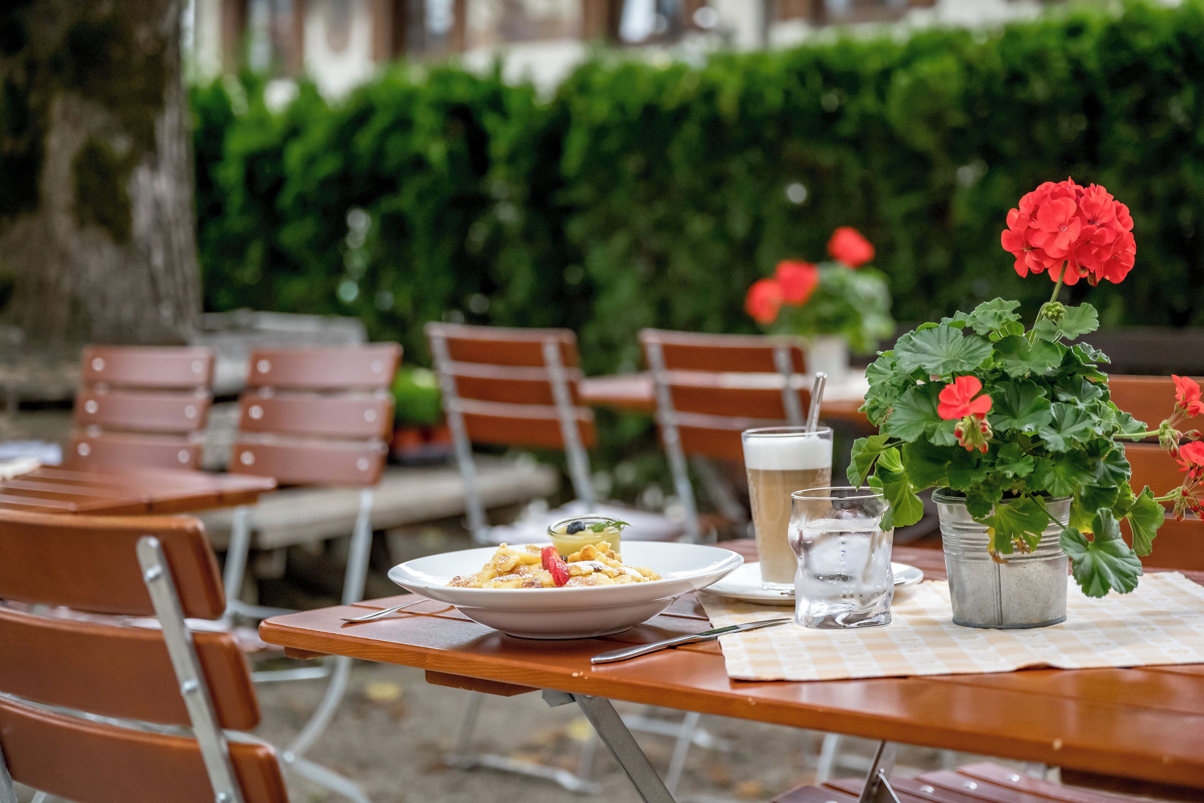 Un espace de restauration en plein air avec une table dressée pour deux personnes, comprenant un plat, un verre de bière et une plante en pot ornée de fleurs rouges.