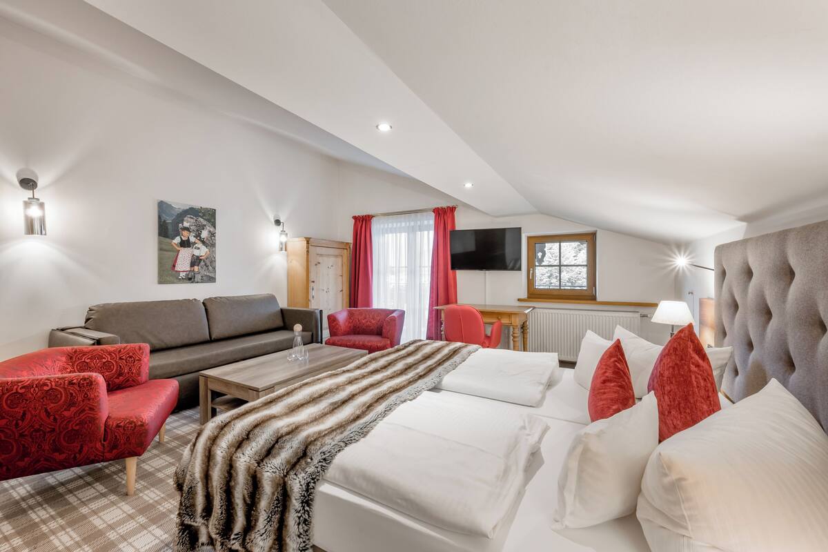 A hotel room with a bed, a sofa, a red armchair, a wooden coffee table, a television, and a window with curtains.