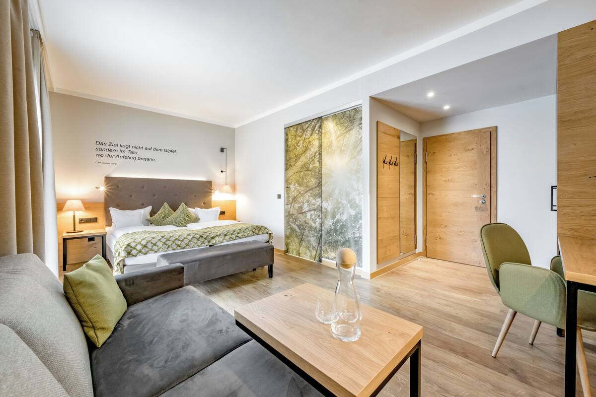 A modern hotel room with a bed, a sofa, a wooden table, and a green chair.