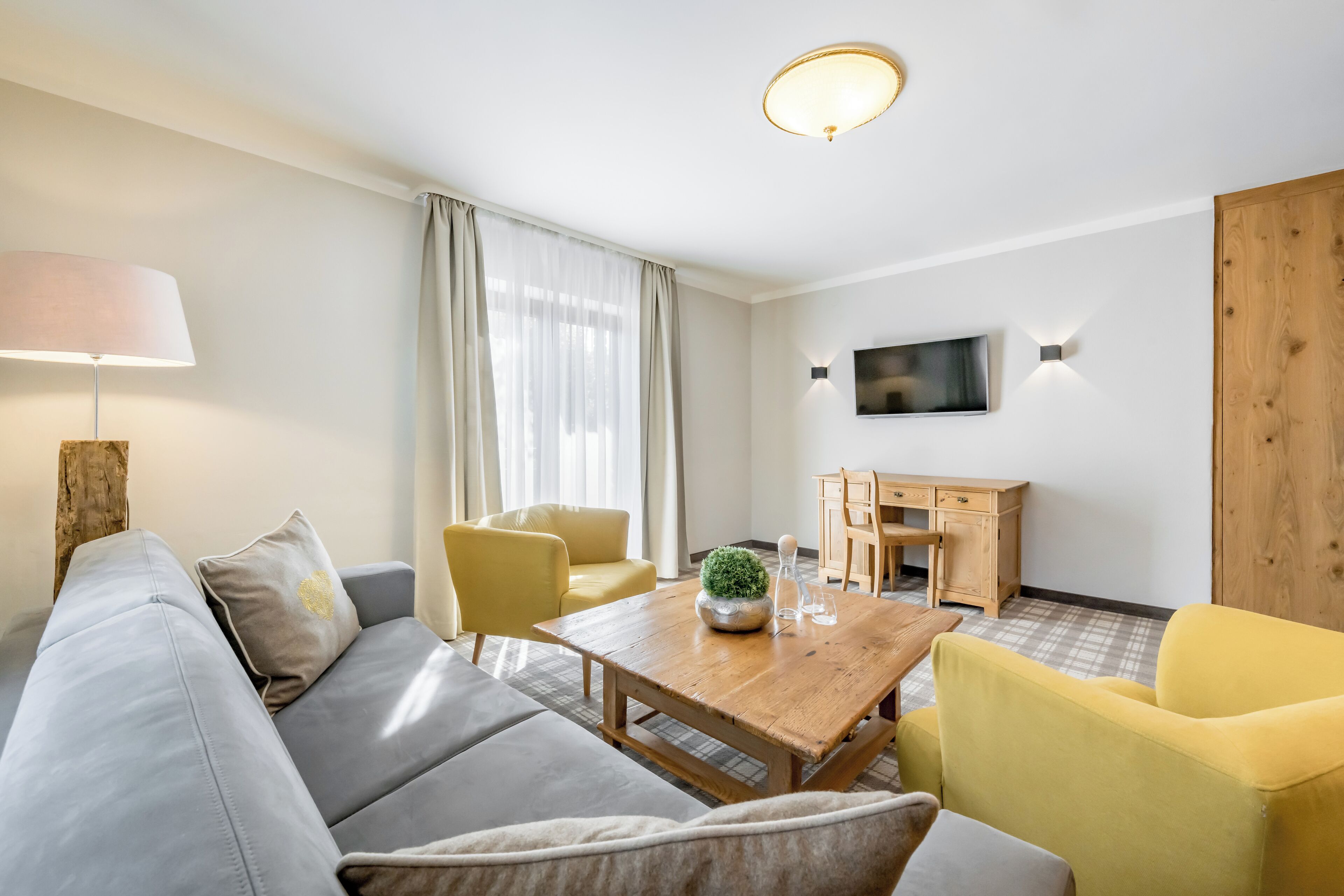 A modern living room with a sofa, yellow armchairs, a wooden coffee table, a TV mounted on the wall, and a wooden cabinet.