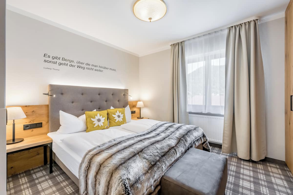 A hotel room with a large bed, a headboard, a bedside table with a lamp, a window with curtains, and a quote on the wall.