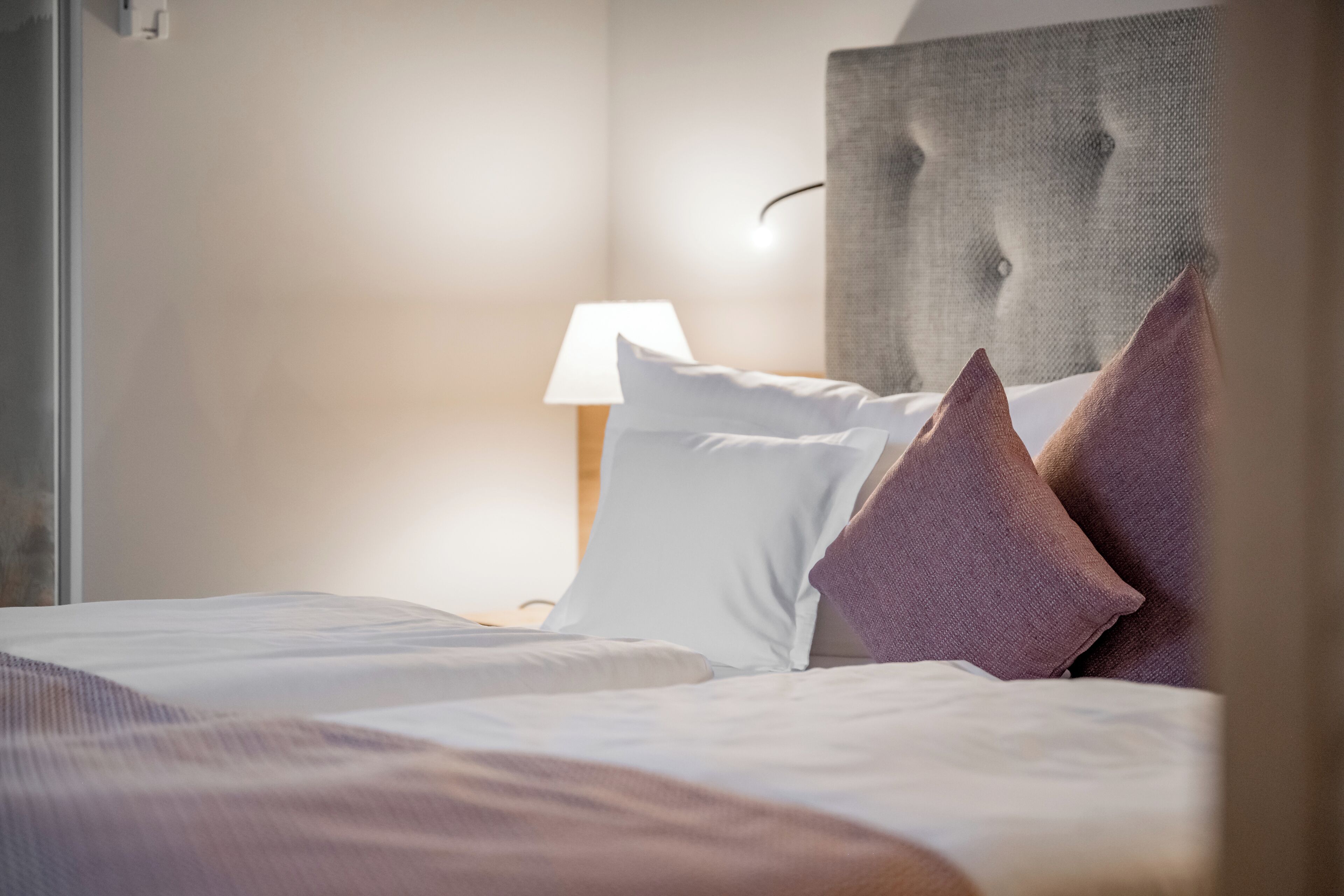 A neatly made bed with white and purple pillows, a lamp on a bedside table, and a textured headboard.