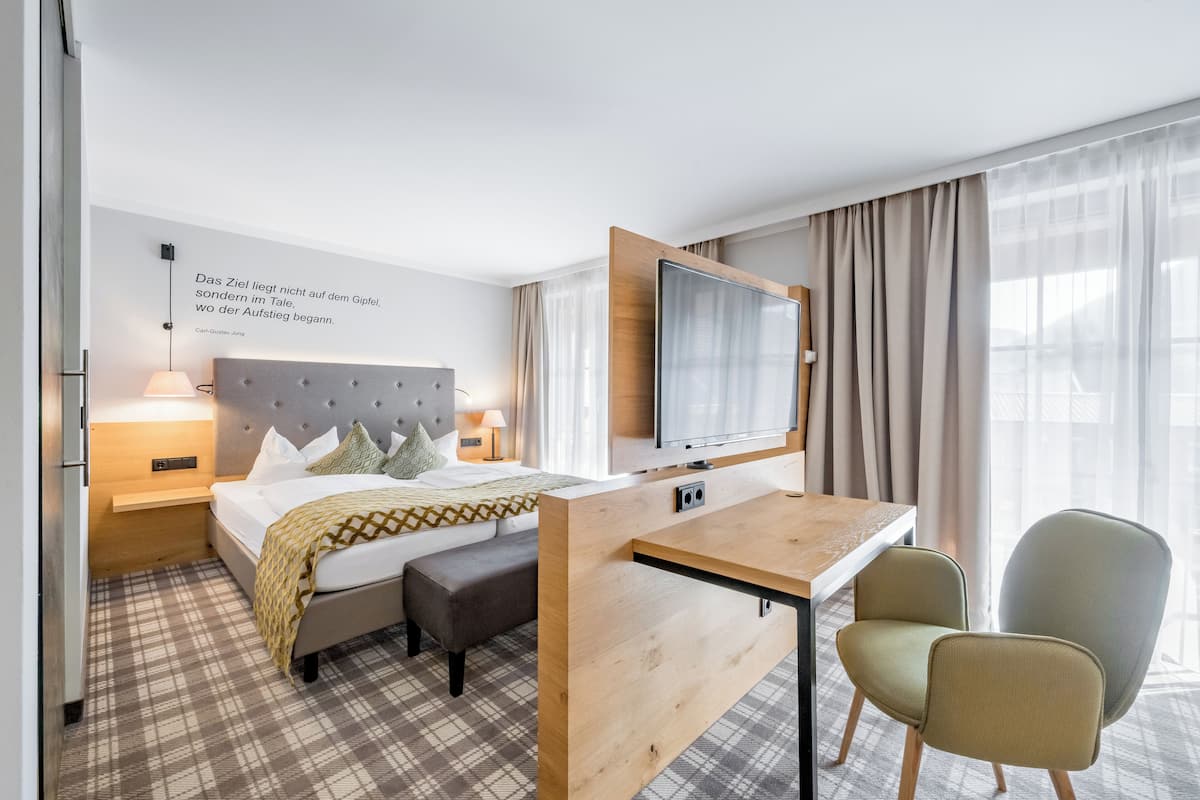 A modern hotel room with a bed, bedside table, lamp, TV, and a desk with chairs.