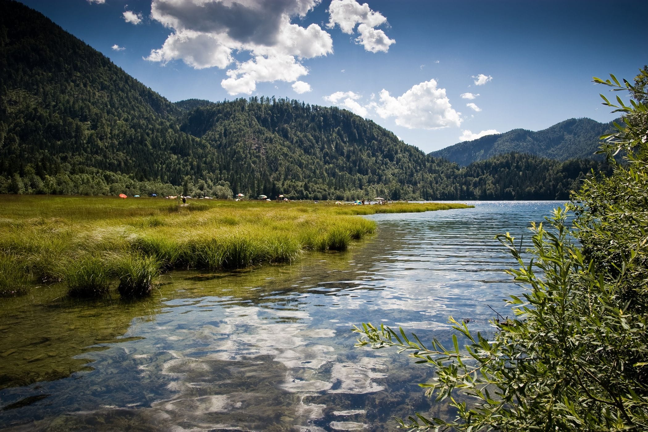 A clear lake surrounded by lush green hills and a few buildings in the distance.