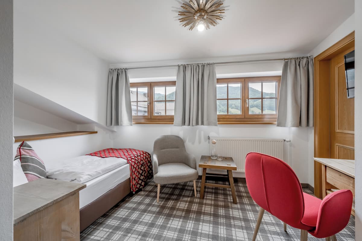A compact room with a bed, a red chair, a small table, and a window with curtains.