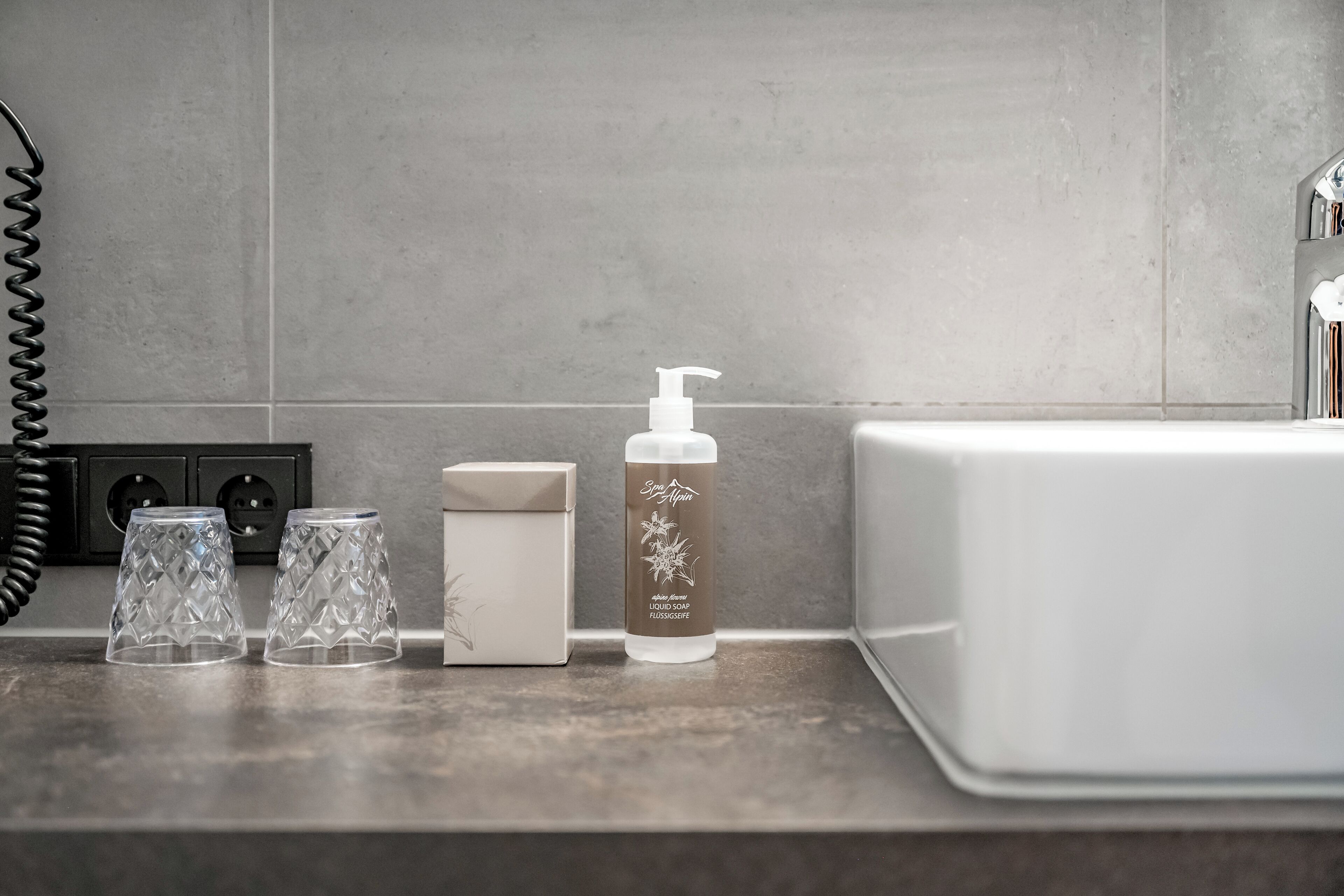 Una encimera de baño con una botella de jabón líquido, dos vasos de vidrio y un jabonera.