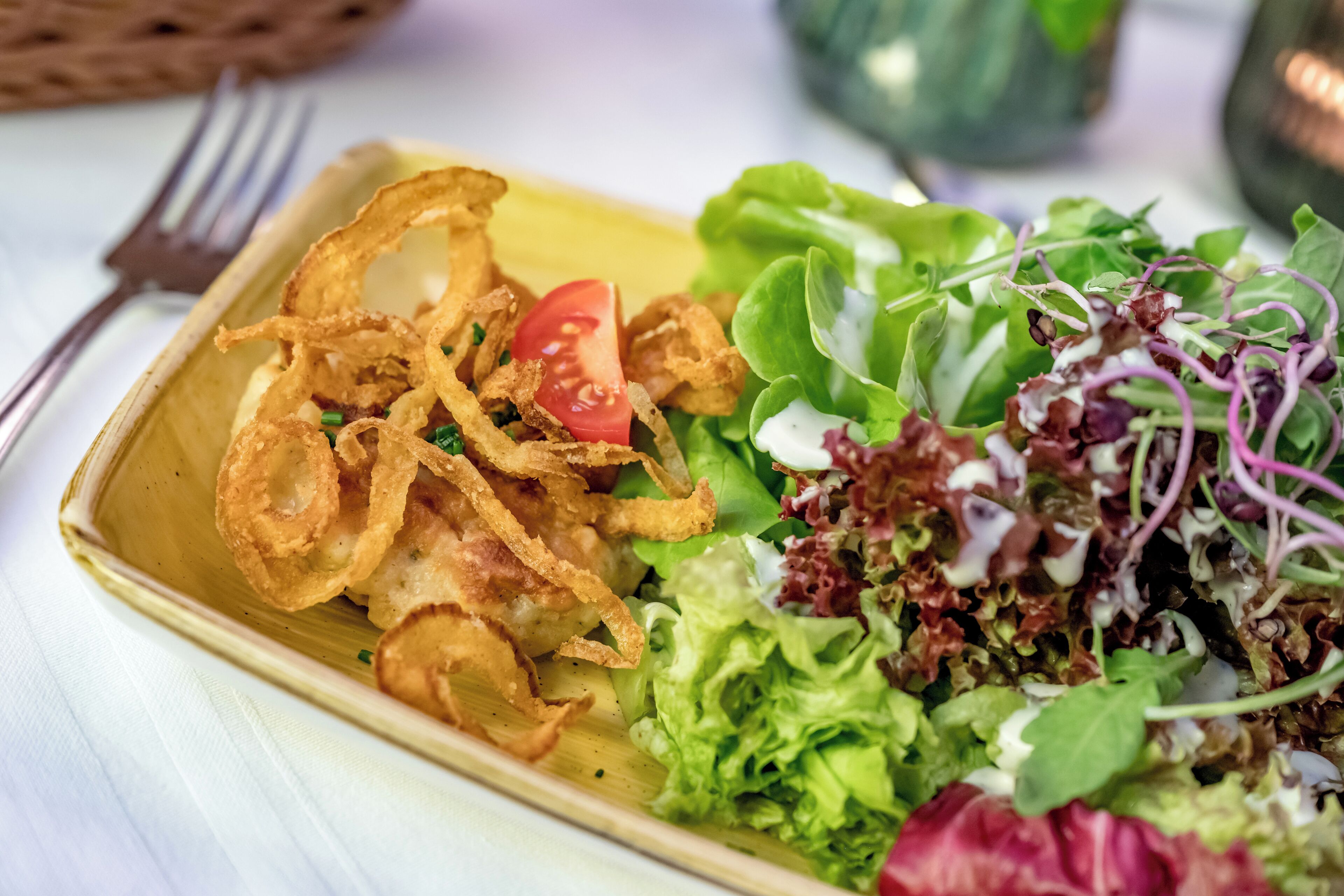 Un plato rectangular con una ensalada de lechuga, tomate y posiblemente algunos brotes, acompañado de aros de cebolla fritos.