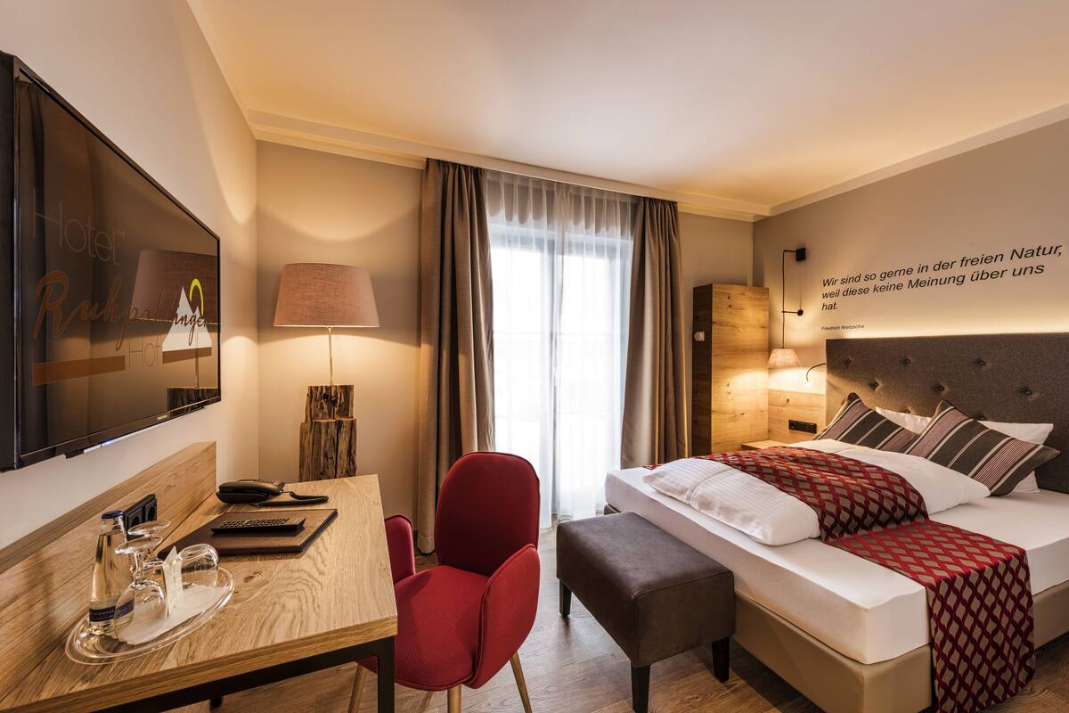 A hotel room with a bed, a desk with a telephone, a chair, a TV, and a window with curtains.
