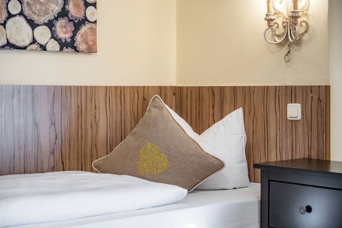A neatly made bed with a wooden headboard, a decorative pillow, a wall-mounted light fixture, and a nightstand.