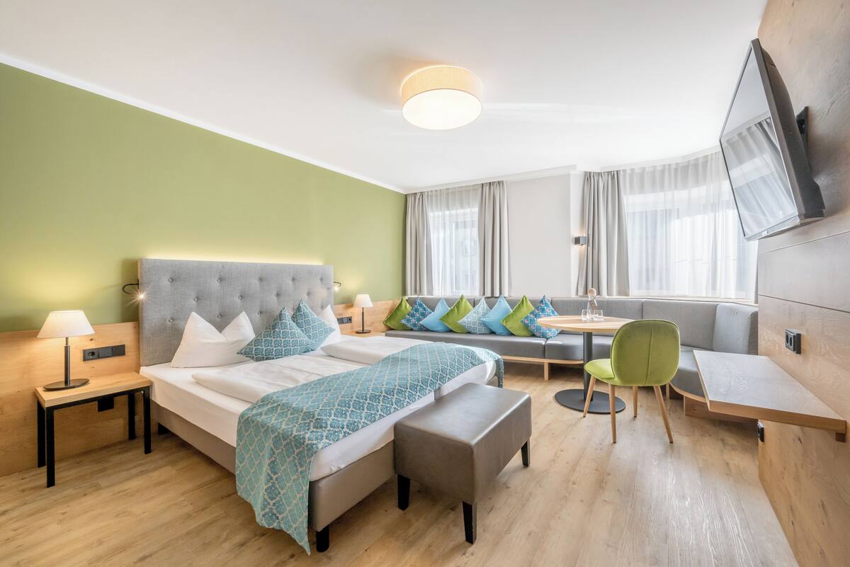 A modern hotel room with a bed, a sofa, a small table, and a TV mounted on the wall.