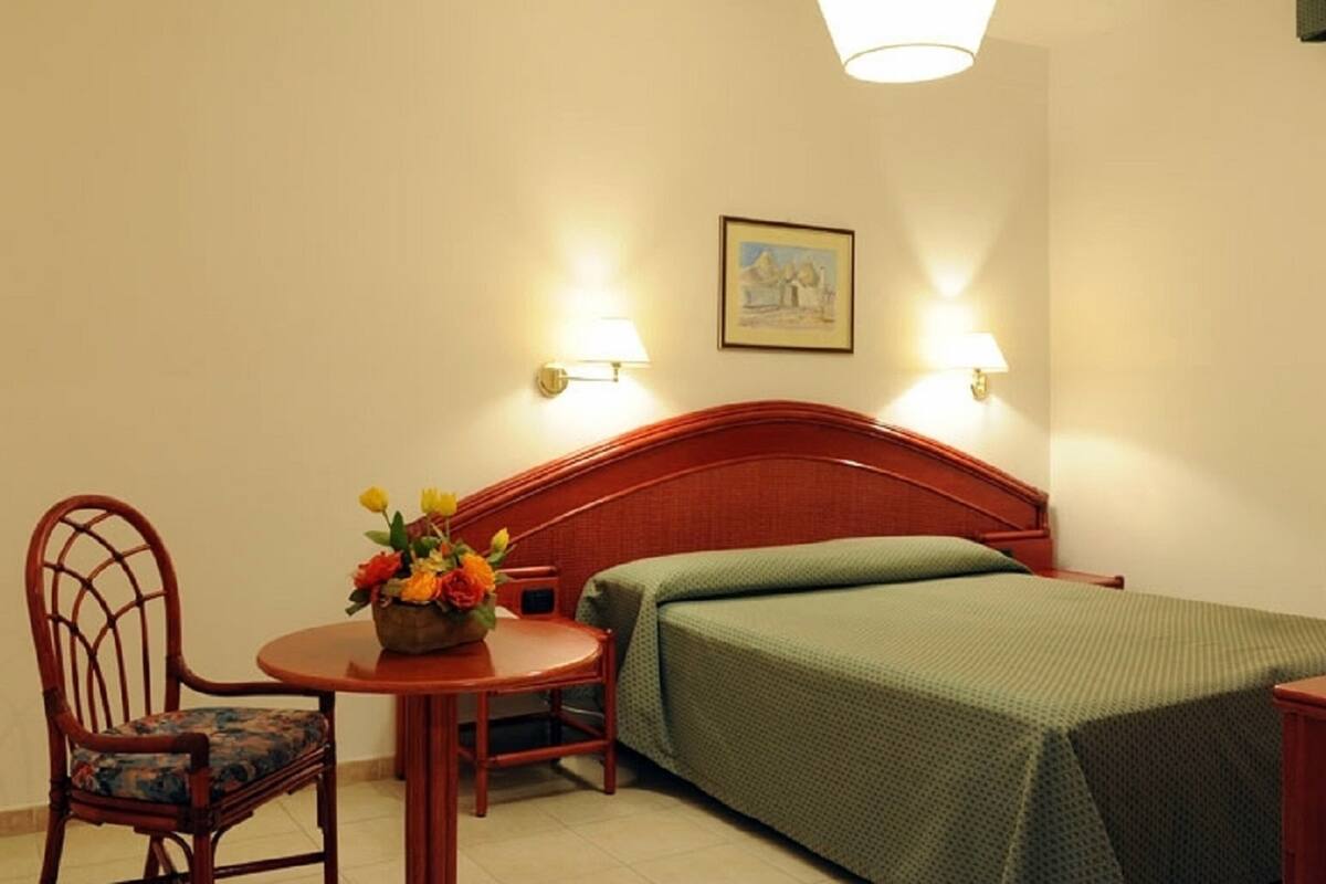 A hotel room with a wooden bed, a green bedspread, a wooden chair with a floral cushion, a small table with a vase of flowers, and two wall-mounted lamps.