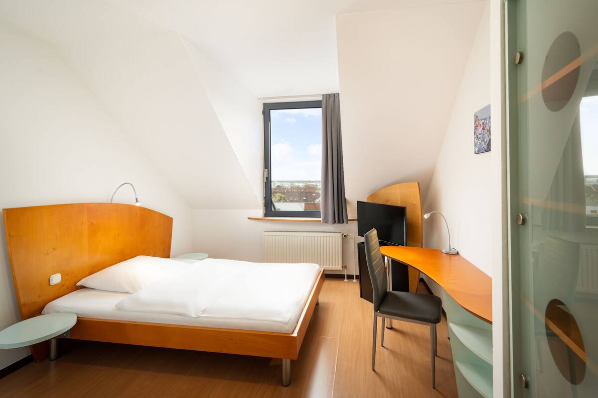 A modern hotel room with a bed, a desk, a chair, a small table, and a window with a view of buildings.