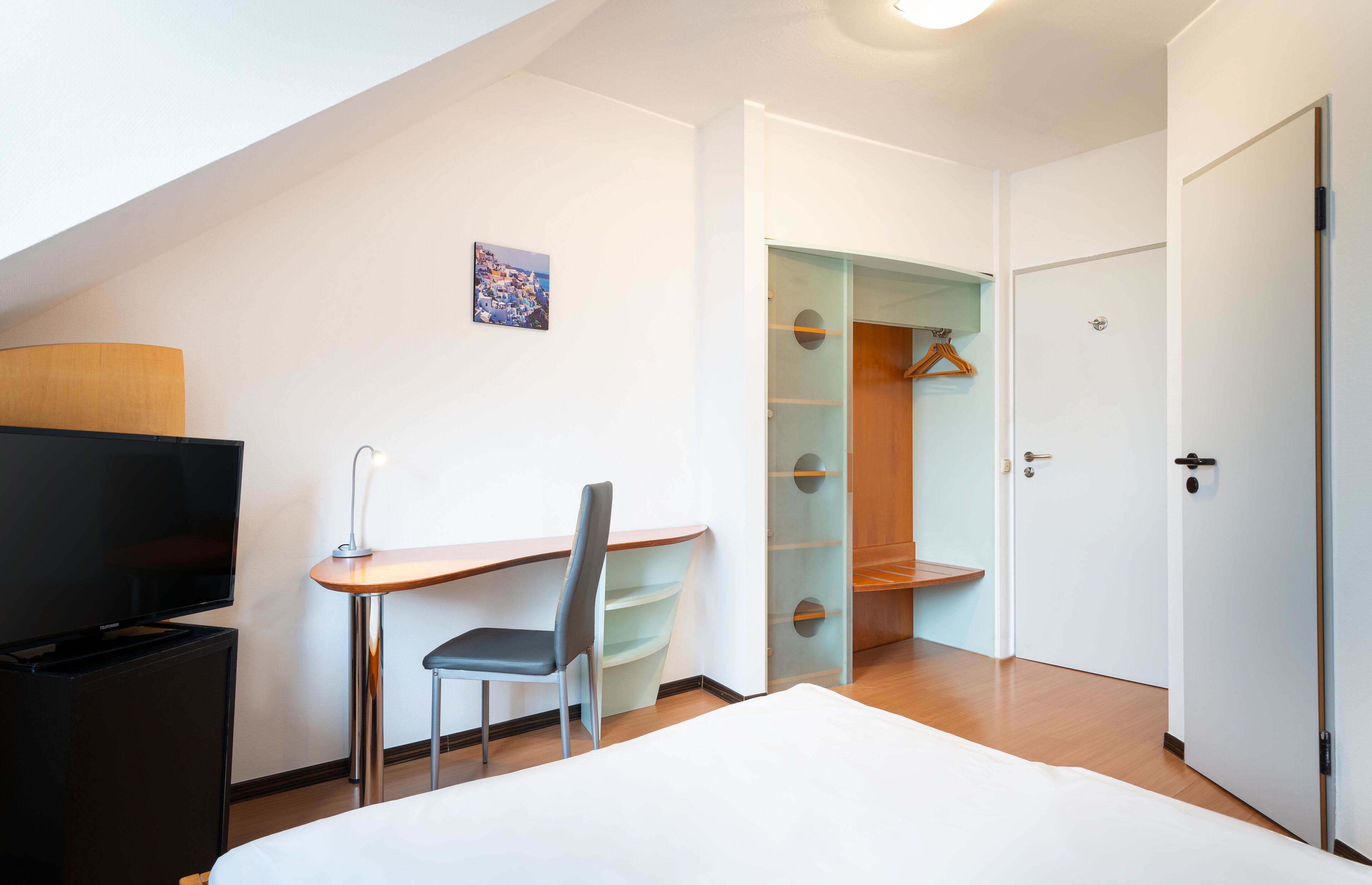 A modern hotel room with a bed, a desk with a chair, a TV, and a walk-in closet.