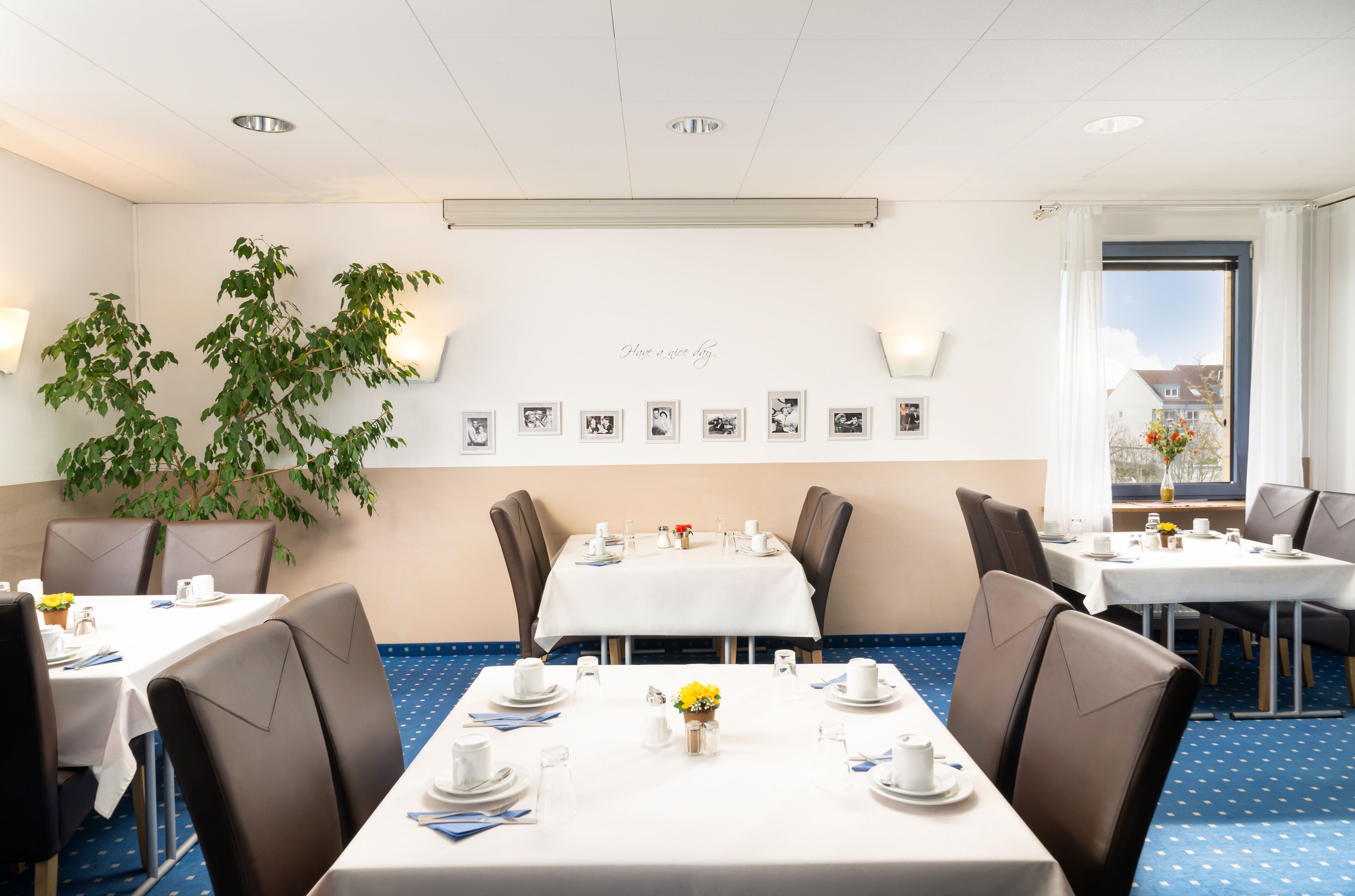 A hotel dining area with tables set for four, a large potted plant, and a wall with framed photos.