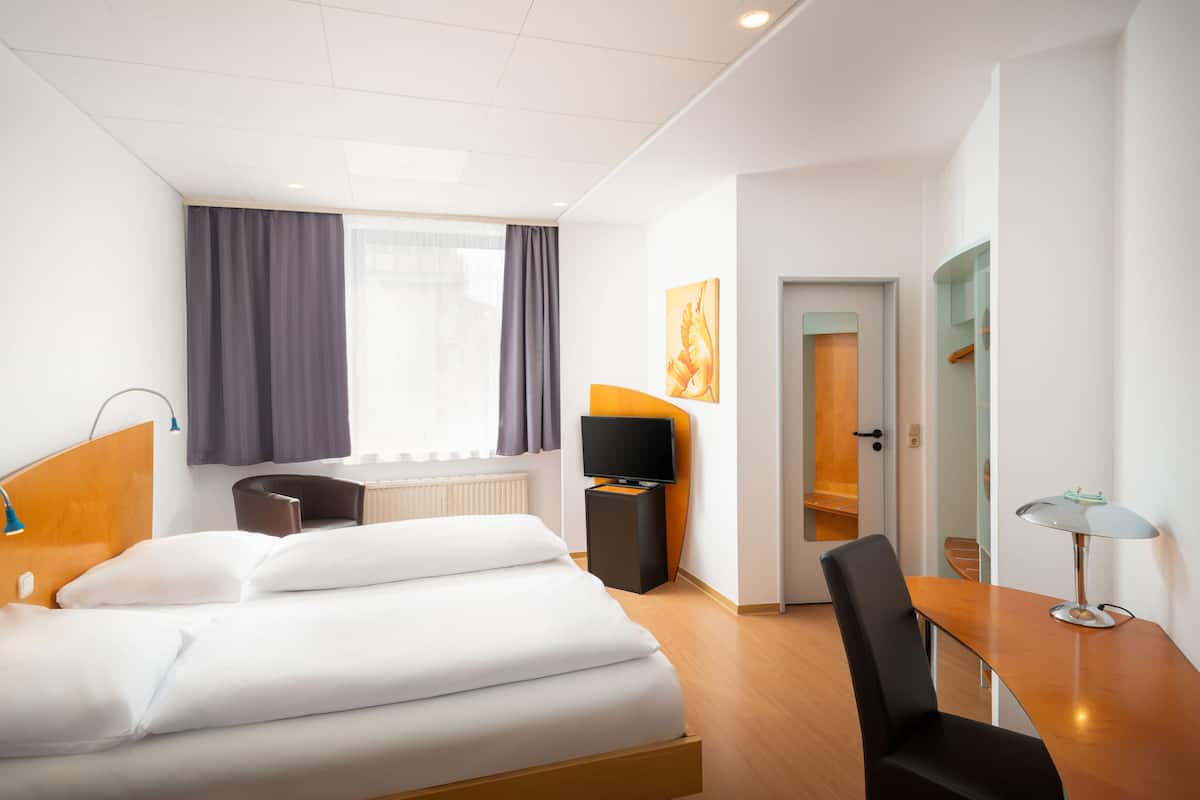 A hotel room with a bed, a desk, a chair, a TV, and a window with curtains.