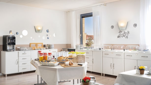 A modern kitchen with white cabinets, a dining table set for breakfast, and a window with curtains. - Hotel Mainstreet (Dietzenbach)