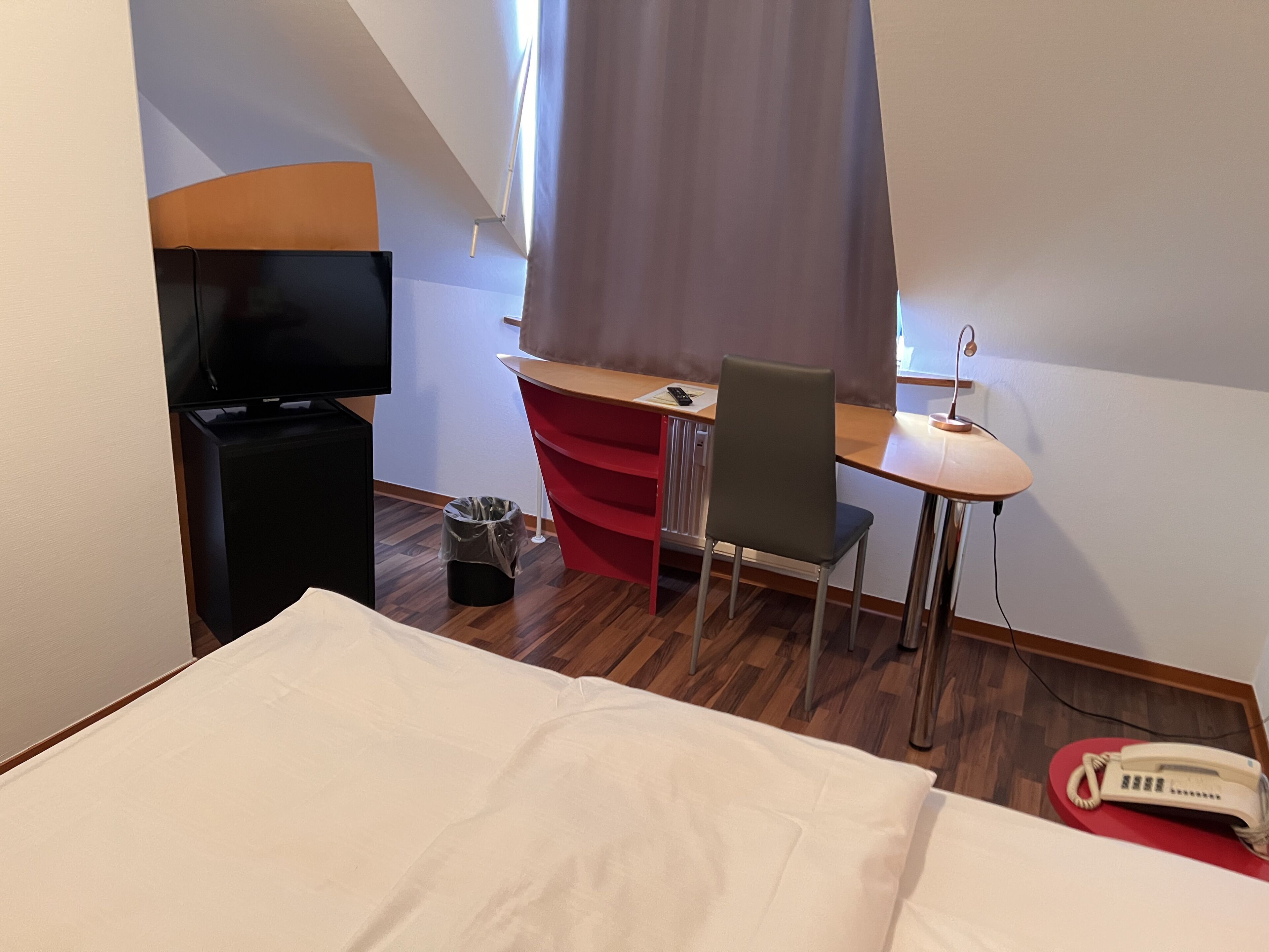 A hotel room with a bed, a desk with a chair, a television, a telephone, and a trash bin.