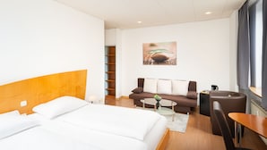 A hotel room with a bed, a sofa, a coffee table, and a chair. There is a painting on the wall and a window with curtains. - Hotel Mainstreet (Dietzenbach)