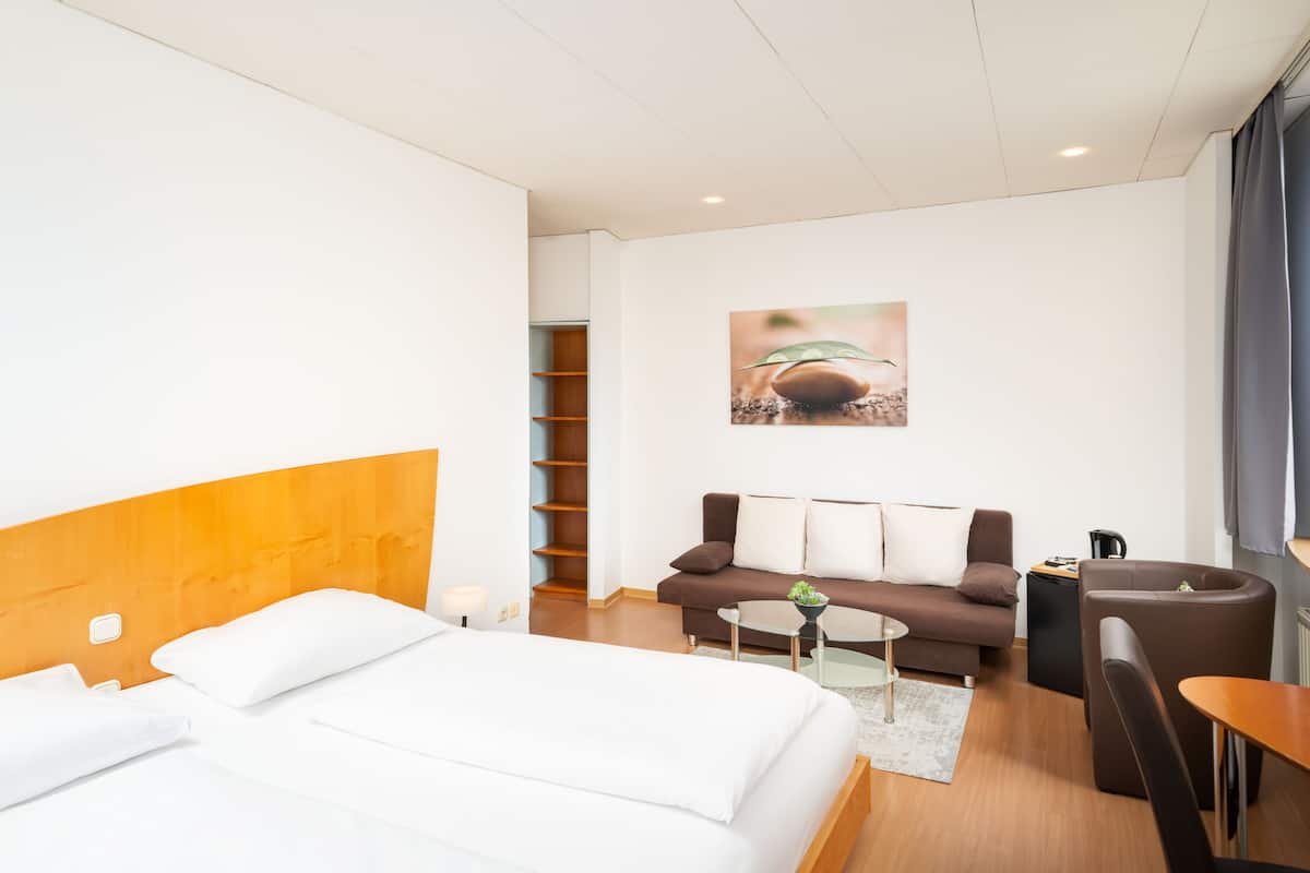 A hotel room with a bed, a sofa, a coffee table, and a chair. There is a painting on the wall and a window with curtains.
