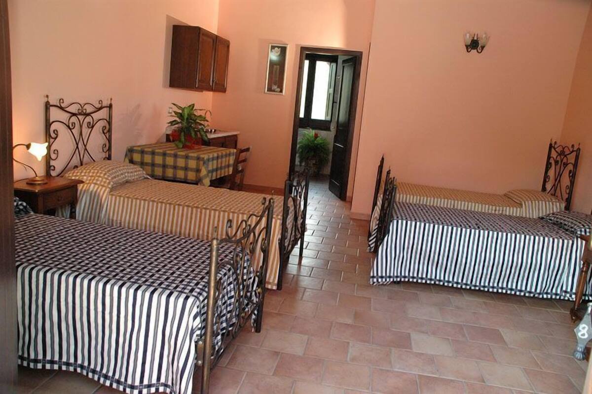 Ein Zimmer mit drei Betten, einem Esstisch mit Stühlen, einer Zimmerpflanze und einer Tür zu einem anderen Raum.