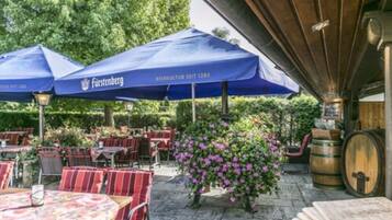 Ein Außenbereich mit Terrasse, Tischen, Stühlen und blauen Sonnenschirmen, einem Holzfässchen und einem blühenden Strauch.