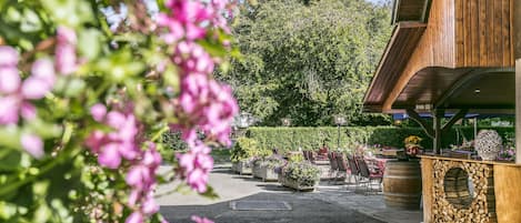 Un espace extérieur aménagé avec des tables et des chaises en bois, entouré d’une végétation luxuriante et de plantes en fleurs.