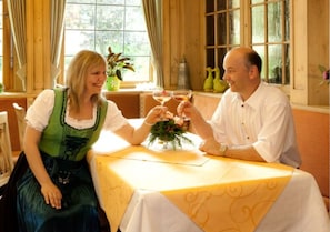 Ein Paar sitzt an einem Tisch mit gelber Tischdecke, ein Fenster mit Vorhängen und ein Blumentopf auf dem Fensterbrett.
