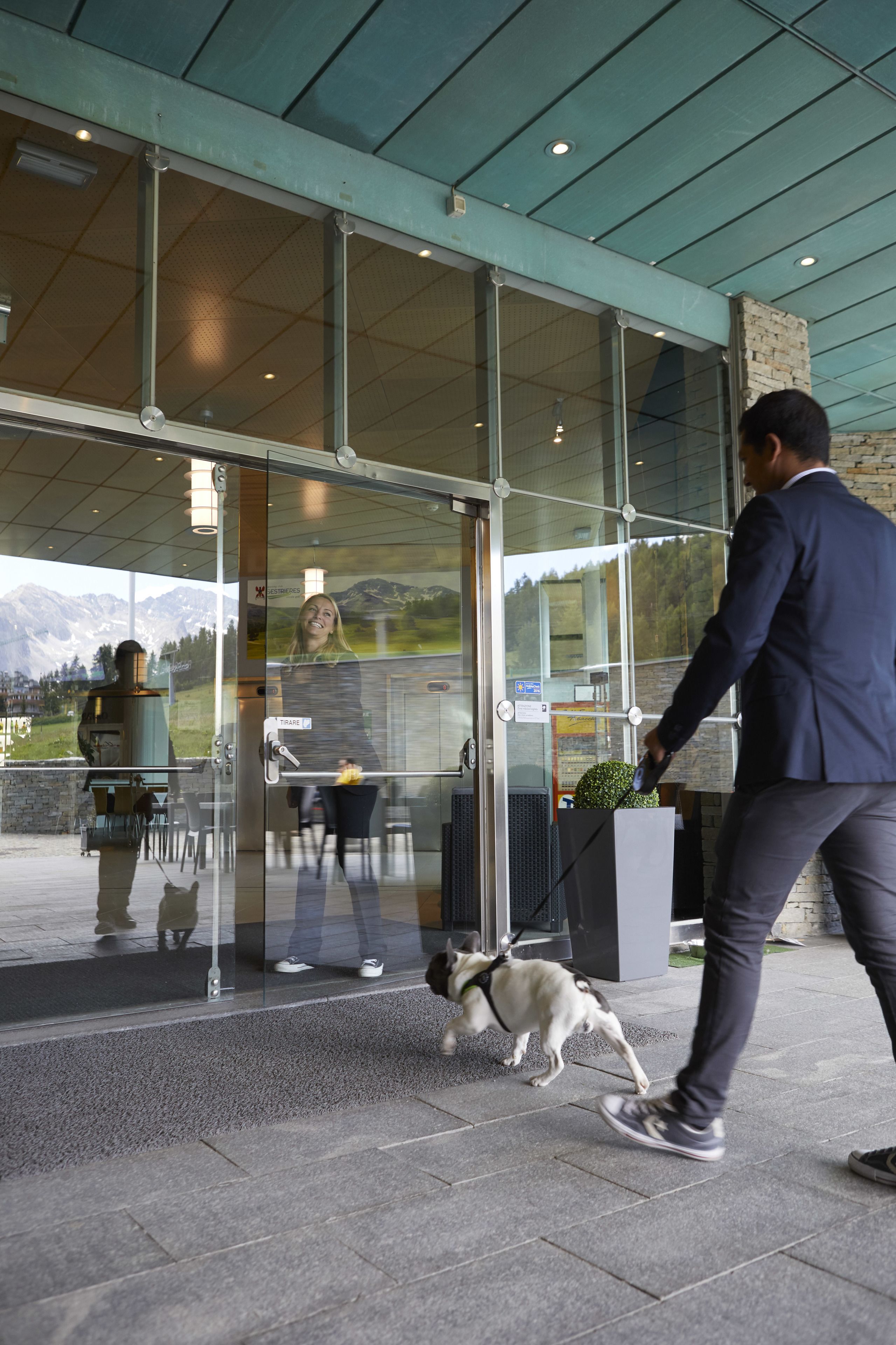 A modern building entrance with glass doors, a stone wall, and a view of mountains in the background.