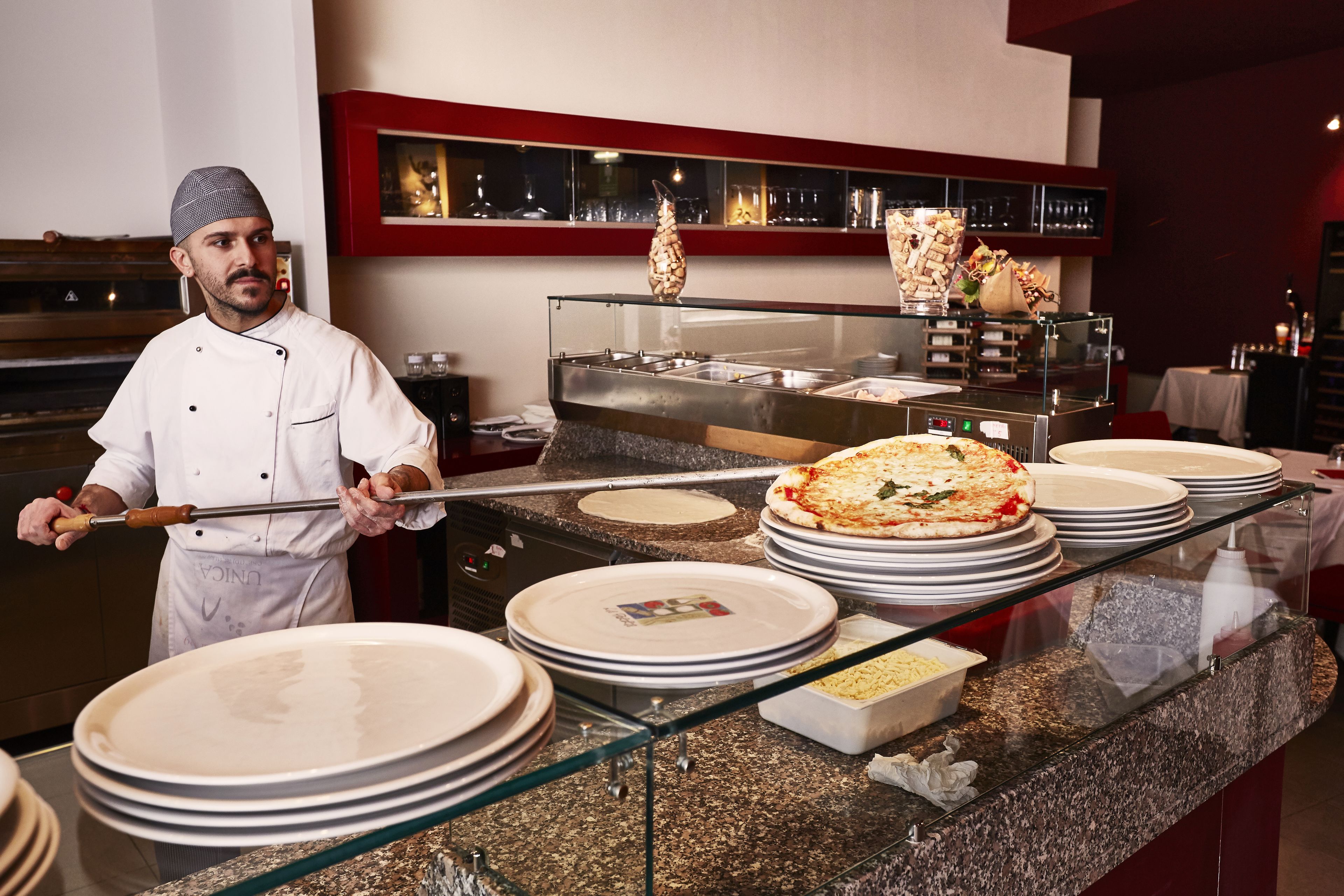 A chef in a kitchen with pizza on display, plates stacked, and a buffet area with food containers.