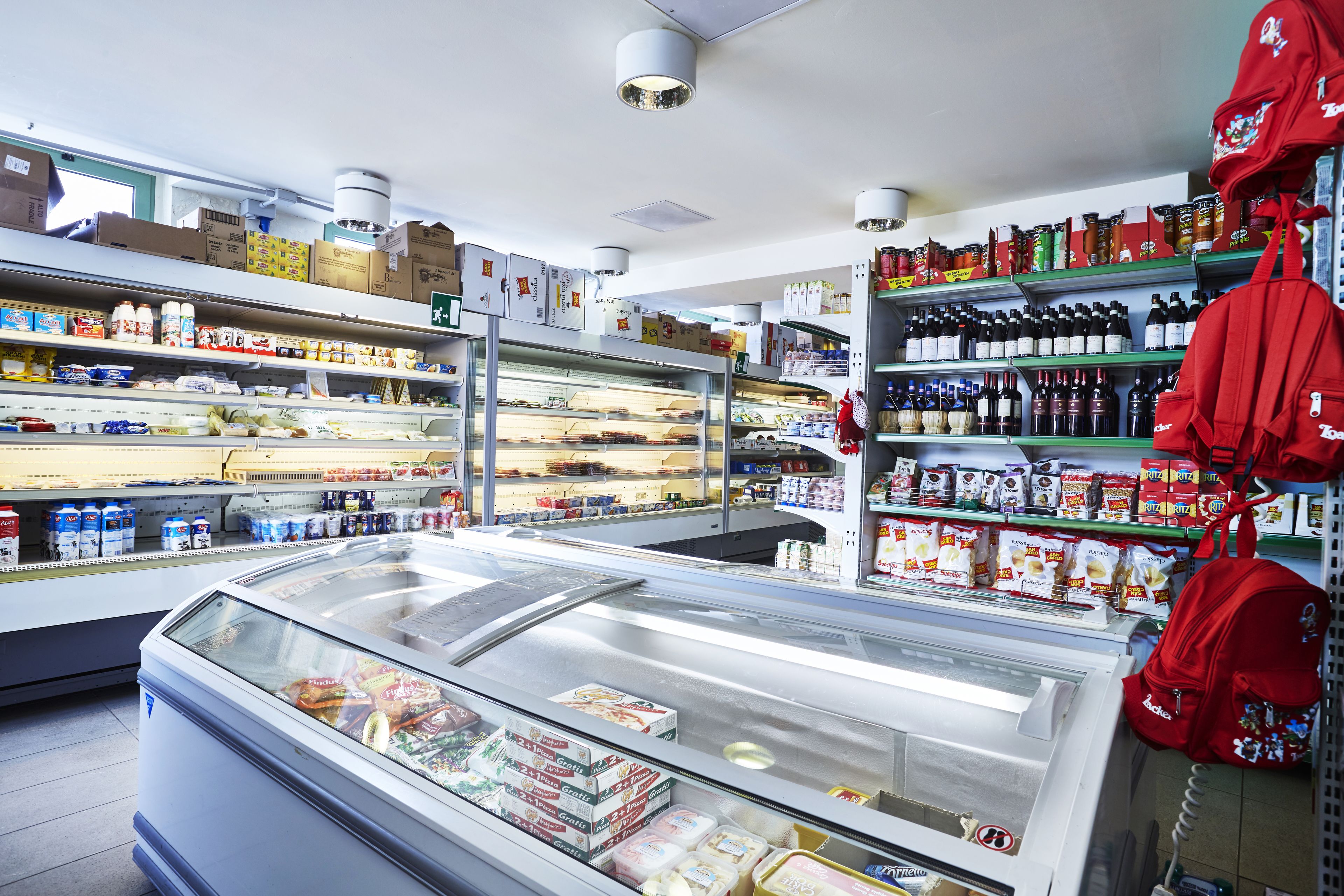 Un supermercado con secciones refrigeradas, estantes llenos de diversos productos, y una mochila roja colgada en la pared.
