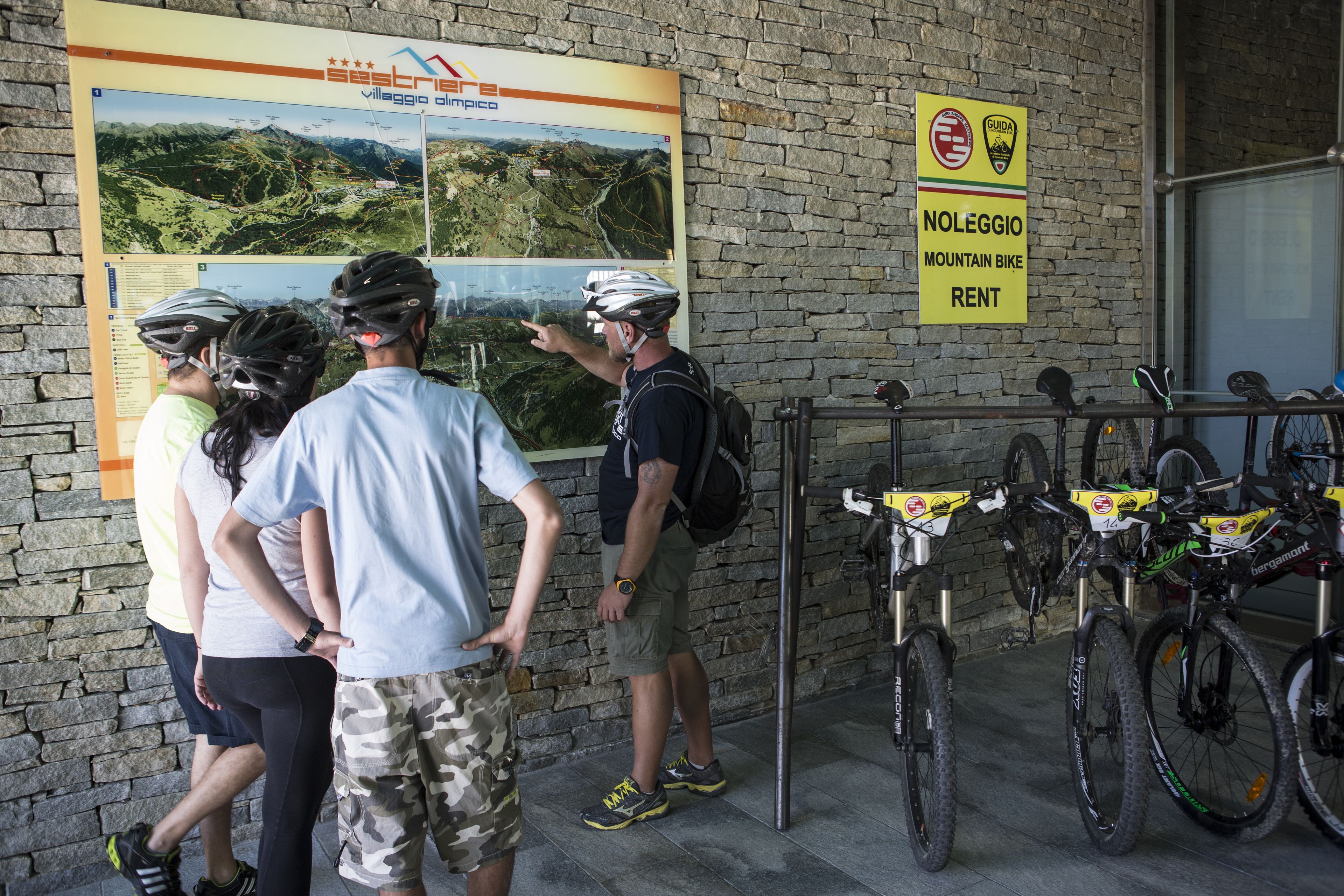 Un grupo de personas en una estación de alquiler de bicicletas de montaña, con un cartel que dice 'Noleggio Mountain Bike Rent' y un mapa en la pared.