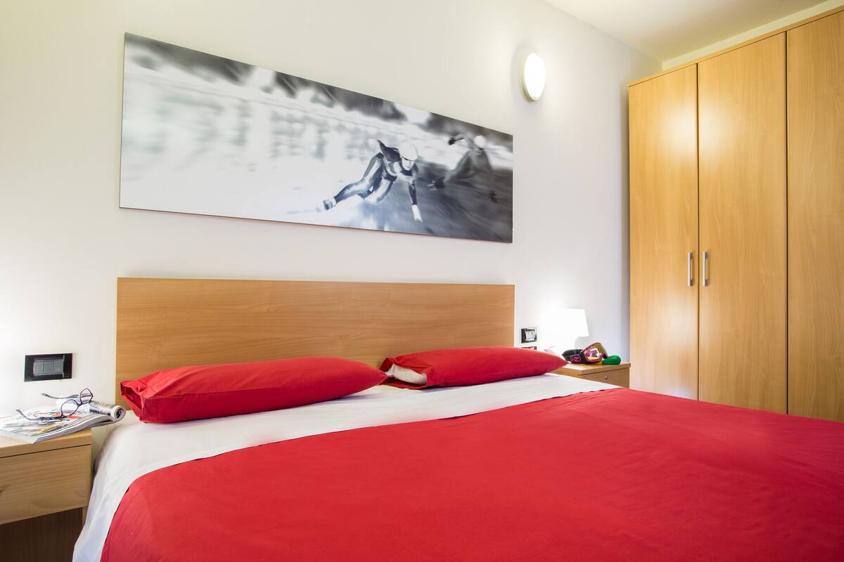 A hotel room with a bed, red bedding, a wooden headboard, a nightstand with a lamp, and a wardrobe.
