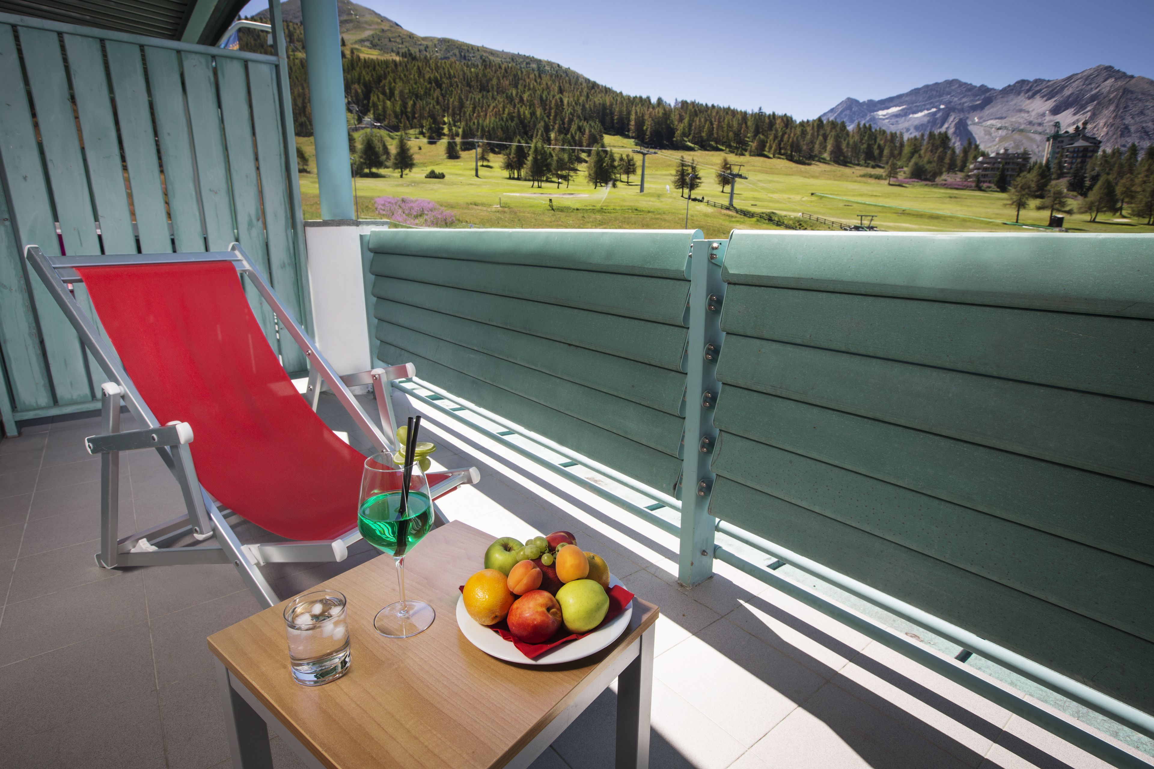 A balcony with a red chair, a table with a fruit plate, a glass of water, and a drink.