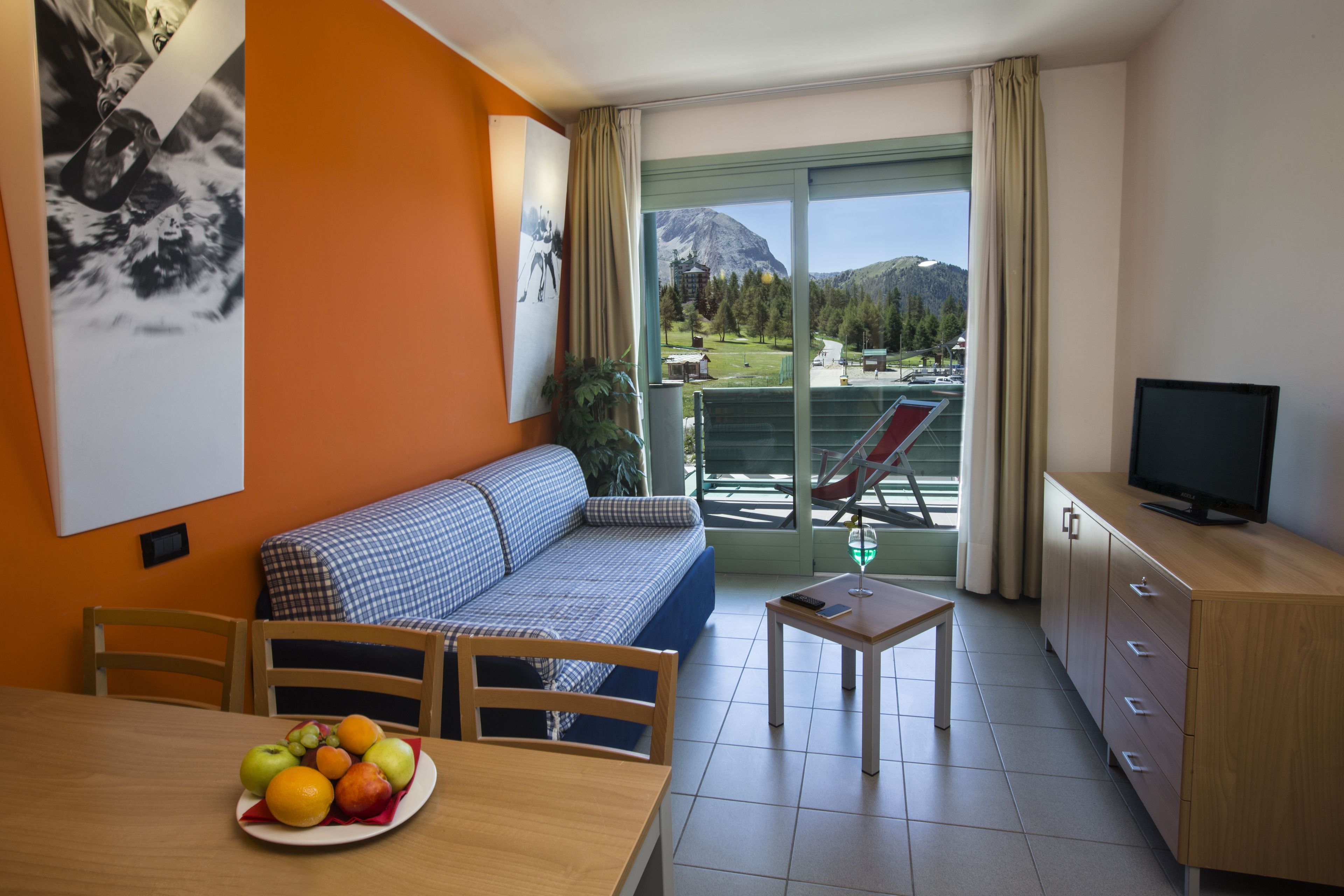 A hotel room with a balcony, a sofa, a dining table with fruit, a TV, and a view of mountains.