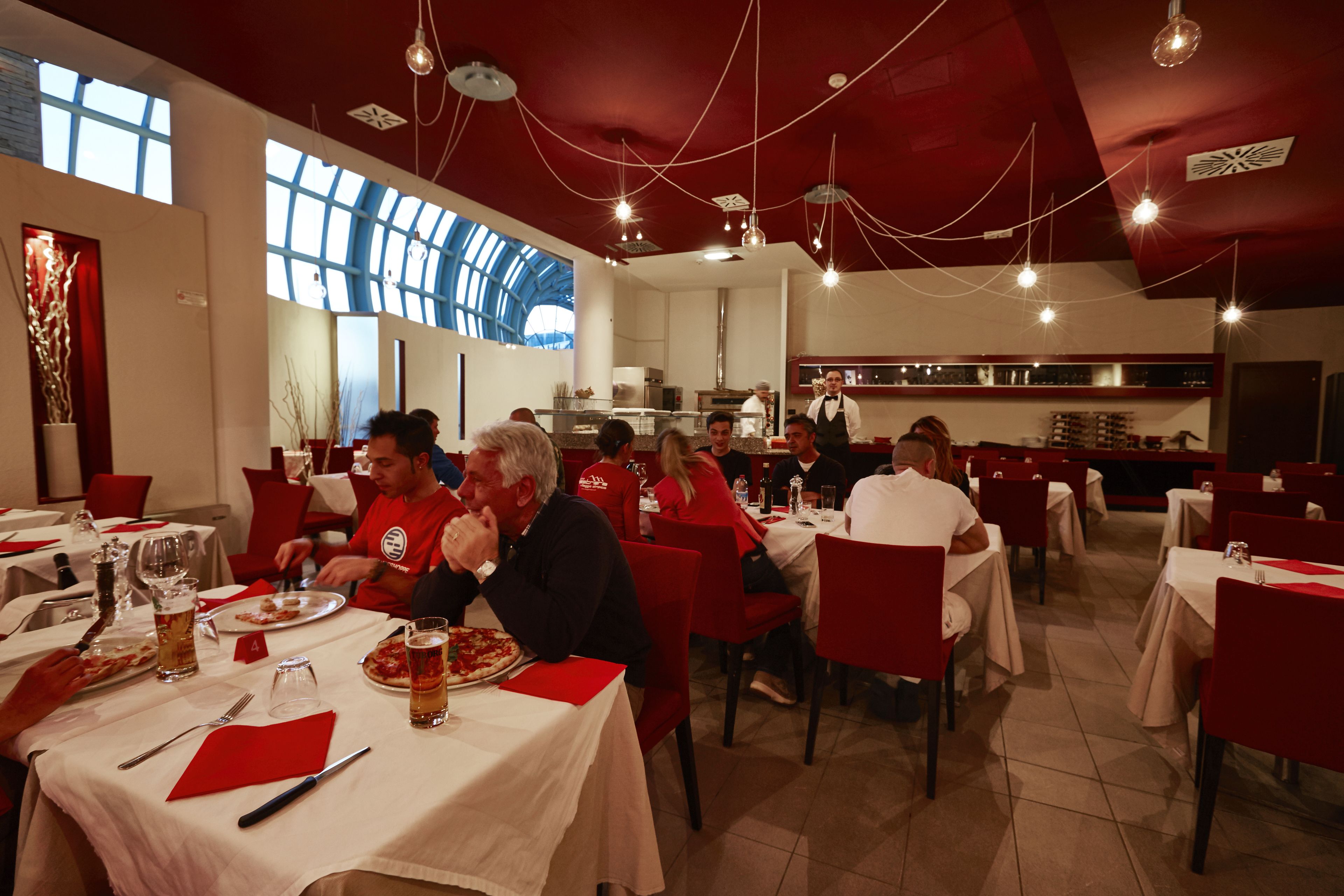 Un restaurante con sillas rojas y manteles blancos, ventanas amplias y lámparas colgantes.