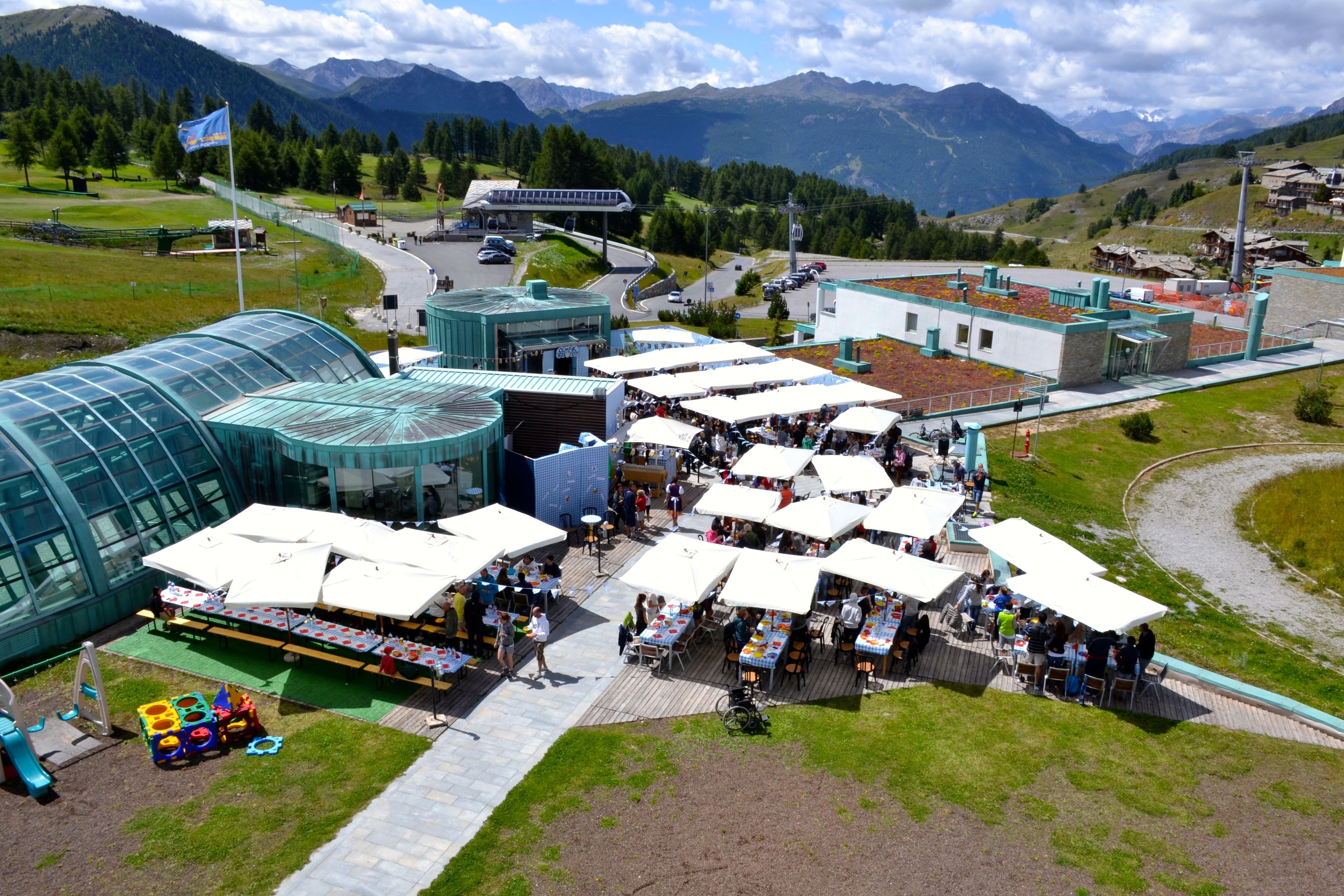 An outdoor market with multiple white umbrellas and tables, surrounded by a mountainous landscape.