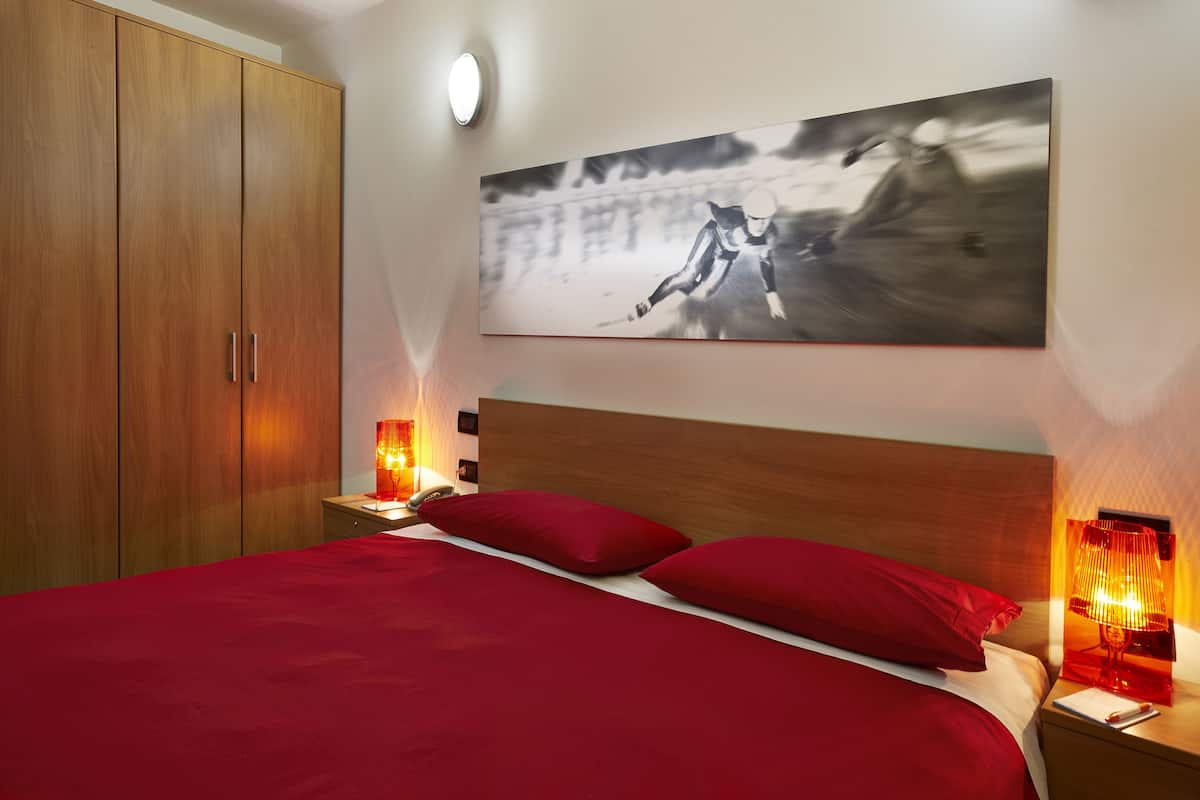 A bedroom with a wooden wardrobe, a bed with red bedding, and two bedside lamps.