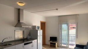 Une cuisine moderne avec des armoires blanches, un évier en acier inoxydable et un four à micro-ondes encastré. On y trouve une petite télévision posée sur un meuble et une porte-fenêtre coulissante donnant sur un balcon.