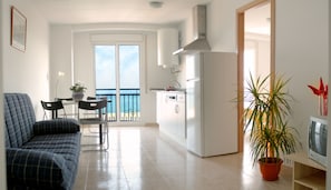 Una sala de estar moderna con un sofá, una mesa de comedor con sillas, una cocina con refrigerador y un balcón con vista al mar.