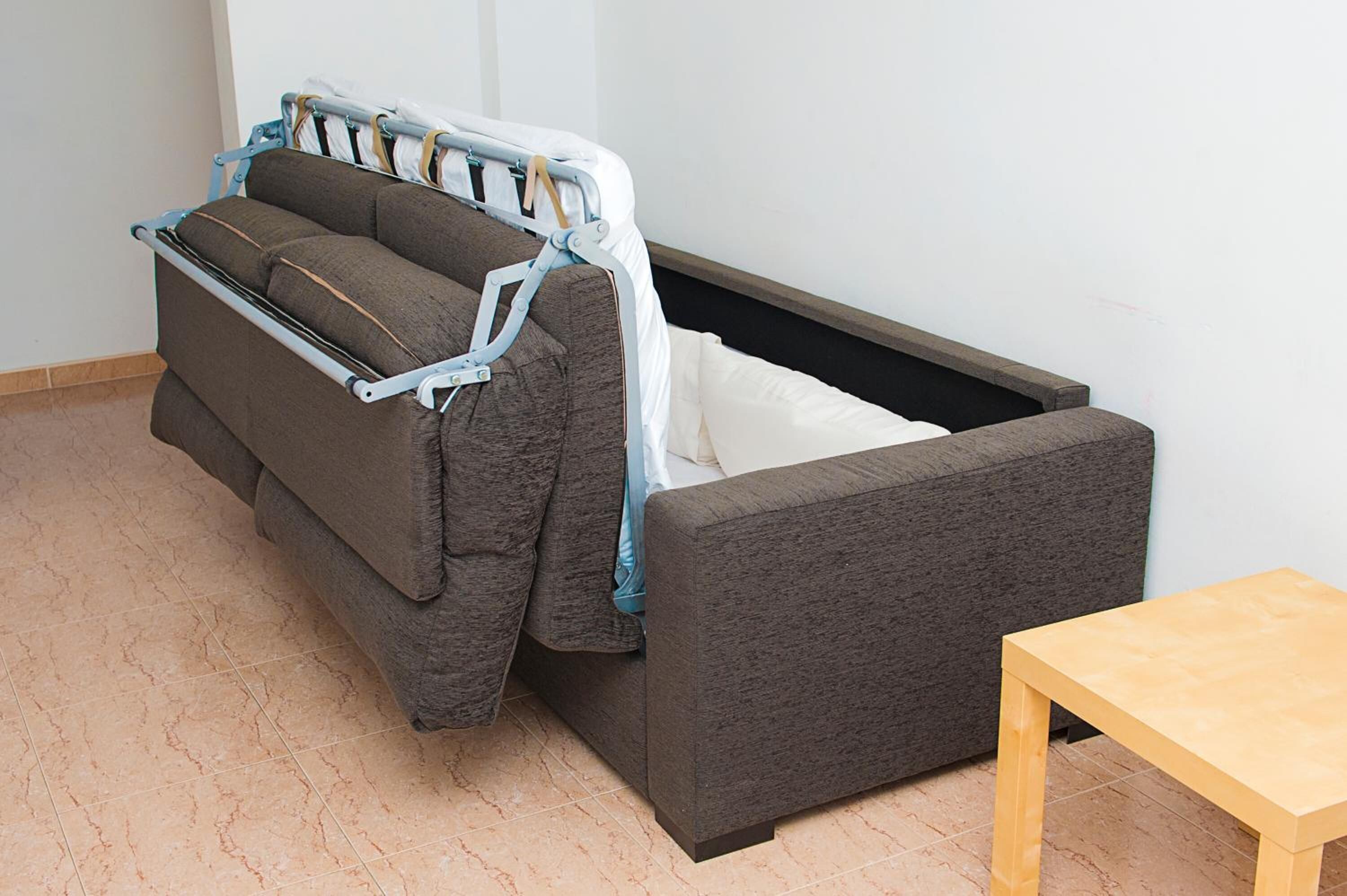 A compact room with a grey sofa bed, a wooden table, and a tiled floor.