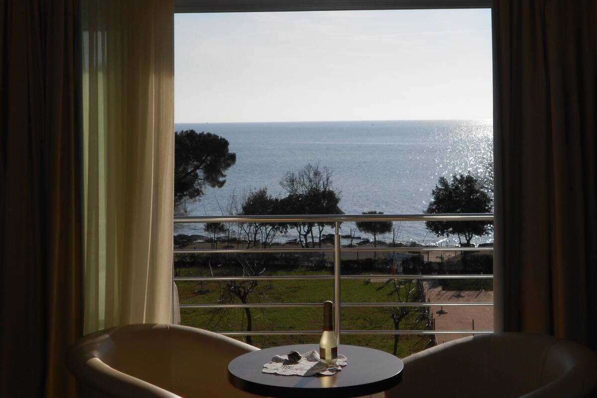 Ein Zimmer mit Meerblick durch ein großes Fenster, ein Tisch mit einer Flasche und zwei Stühle.