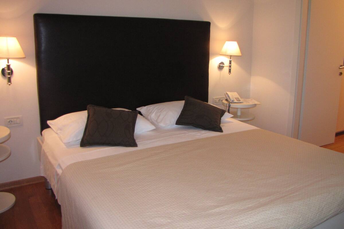 Ein ordentlich bezogenes Bett mit schwarzem Kopfteil, zwei Kissen und einer weißen Tagesdecke. Zwei Nachttischlampen und ein Nachttisch mit Telefon und Lampe sind vorhanden.