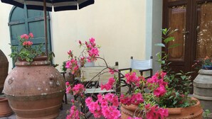 En gårdsplads med store terracotta krukker med lyserøde blomster, en træbænk og en blåstribet parasol.