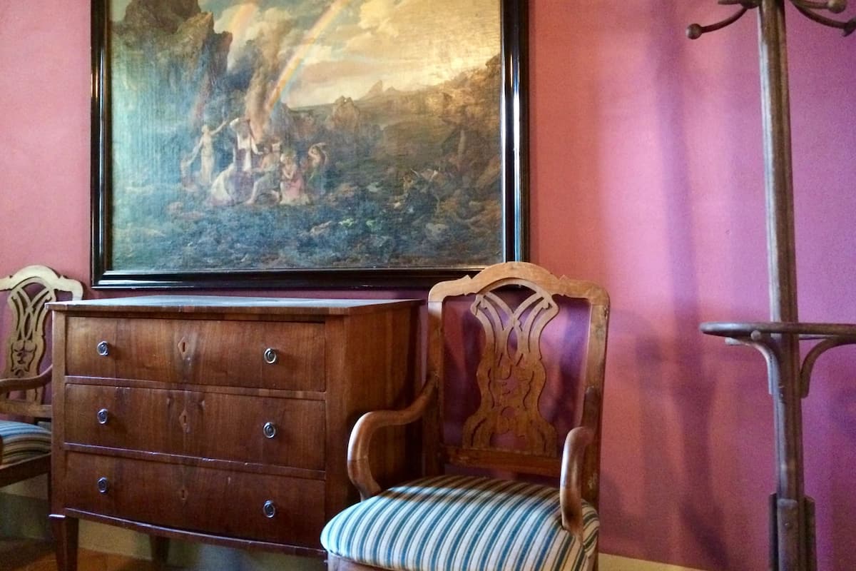 A room with a large landscape painting, a wooden dresser, a striped chair, and a metal stand.